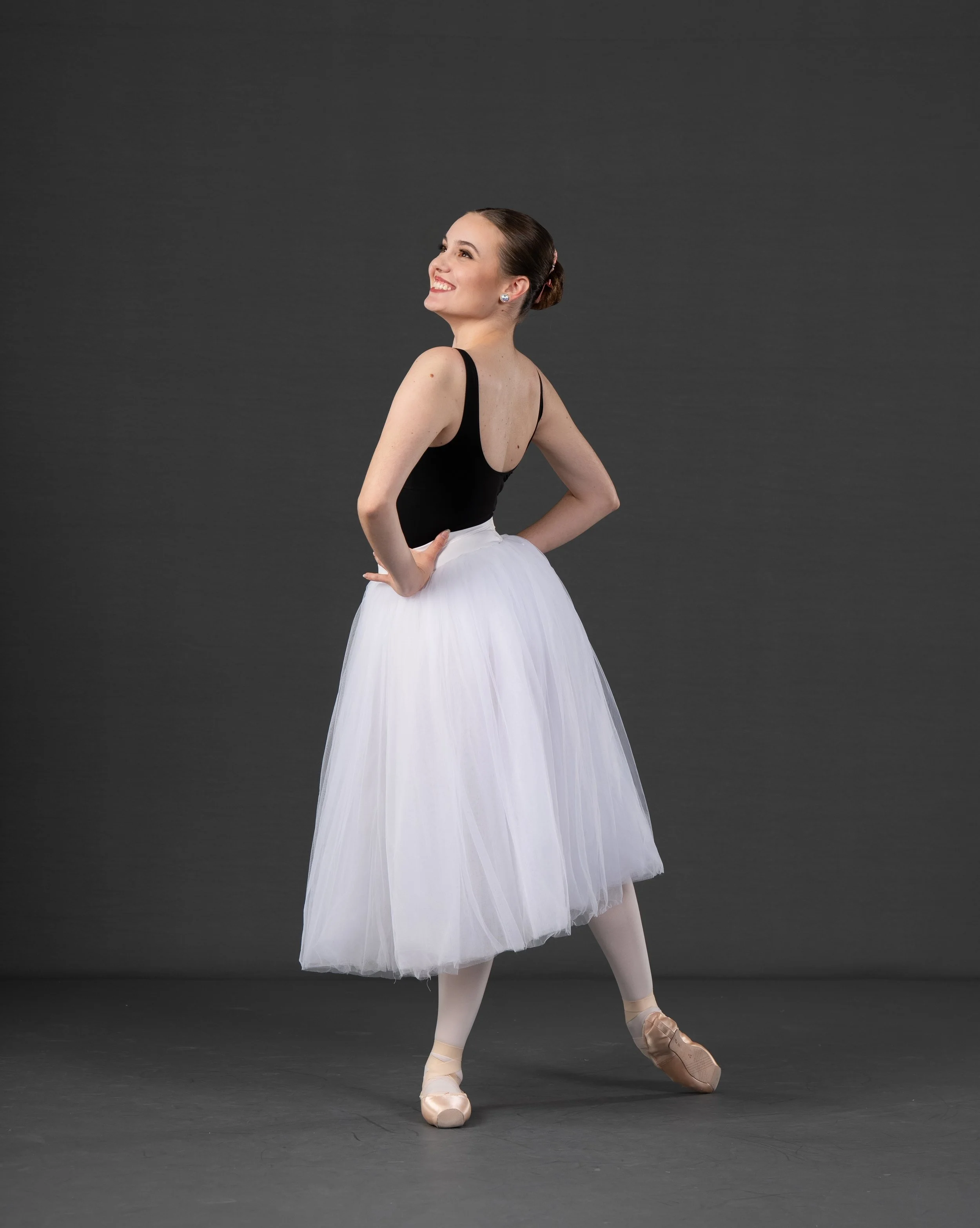 Ballet dancer in a black leotard and white tutu on a dark gray background, standing on tiptoe with one leg bent and arms behind her back.