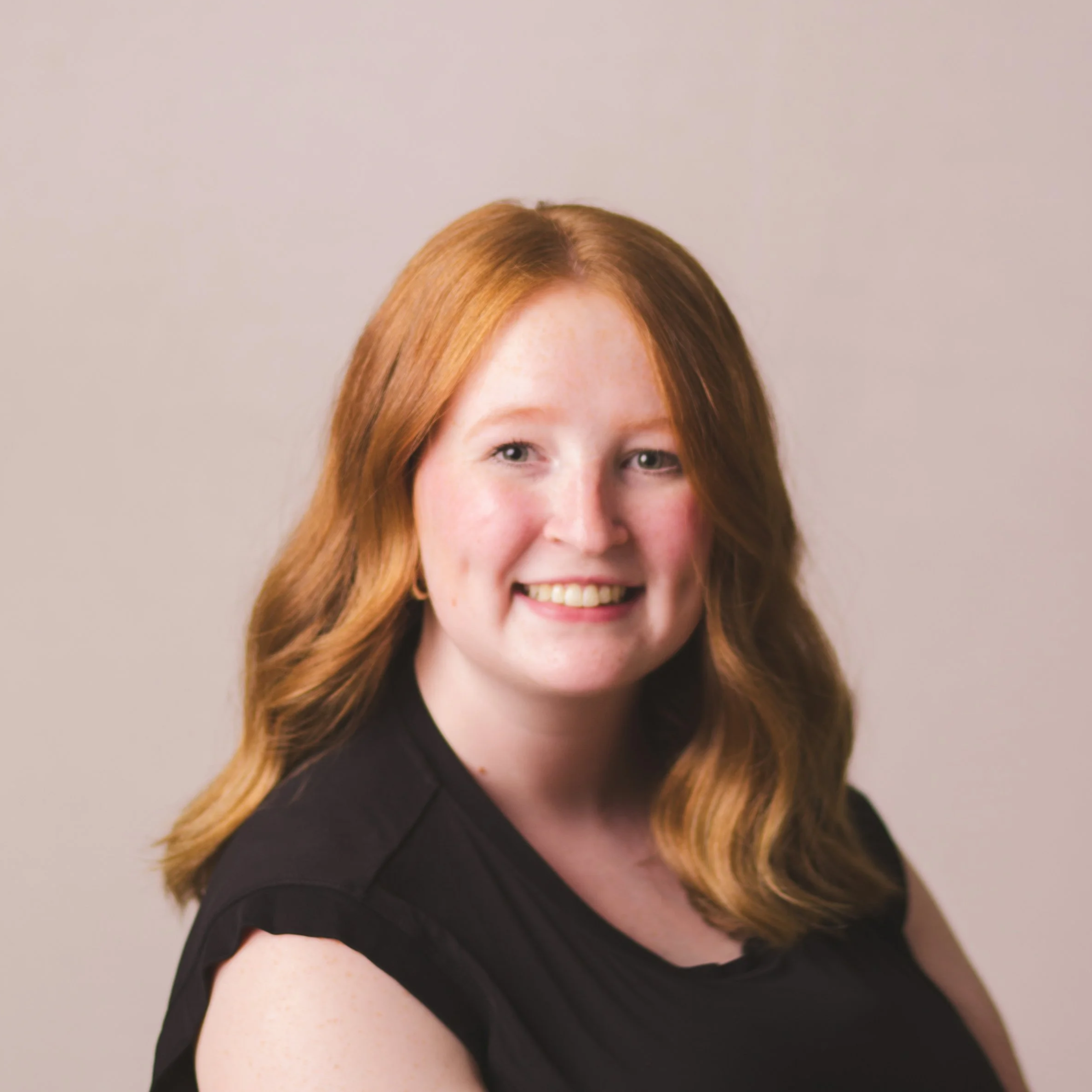 A young woman with wavy, shoulder-length blonde hair, light skin, and blue eyes smiling, wearing a dark gray top, in front of a plain white background.