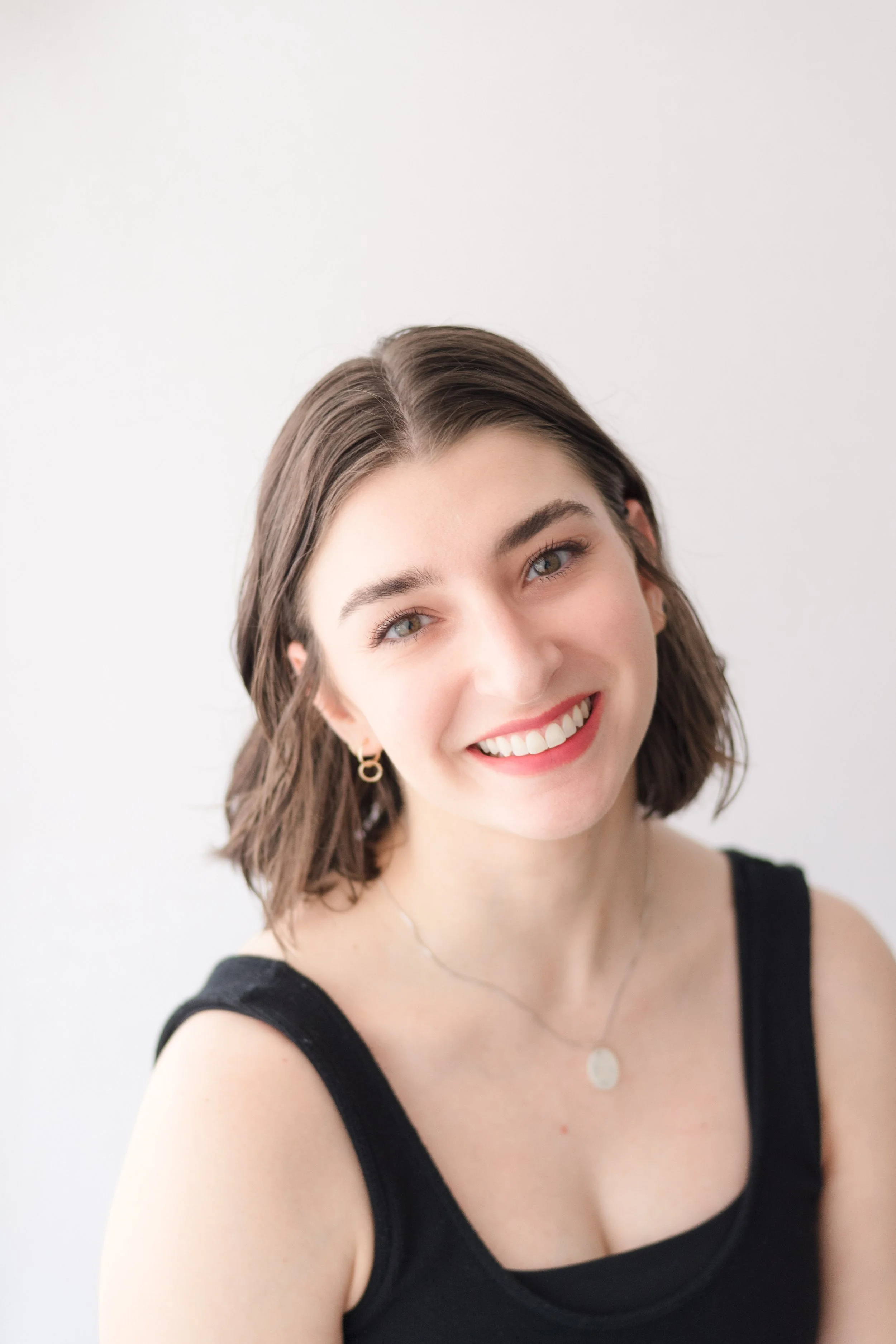 A young woman with shoulder-length brown hair, wearing a black tank top, gold hoop earrings, and a silver necklace, smiling against a plain white background.