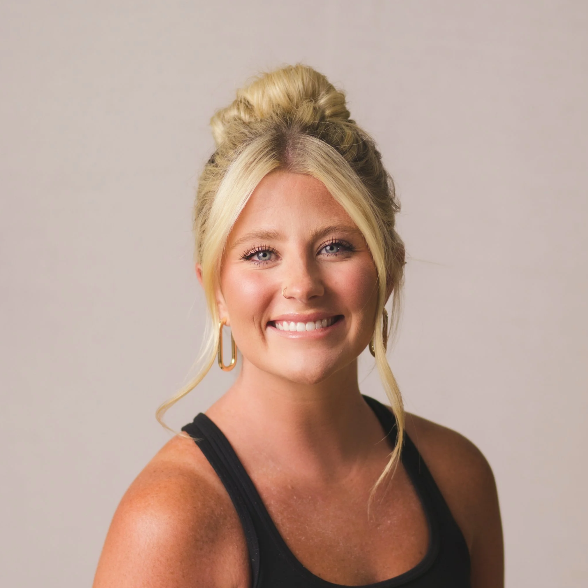 A smiling blonde woman wearing a pink off-the-shoulder ruffled dress and gold jewelry, standing against a light background.