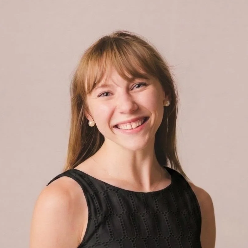 A smiling young woman with short brown hair and blue eyes, wearing a black top and a dangling earring, standing against a gray background.