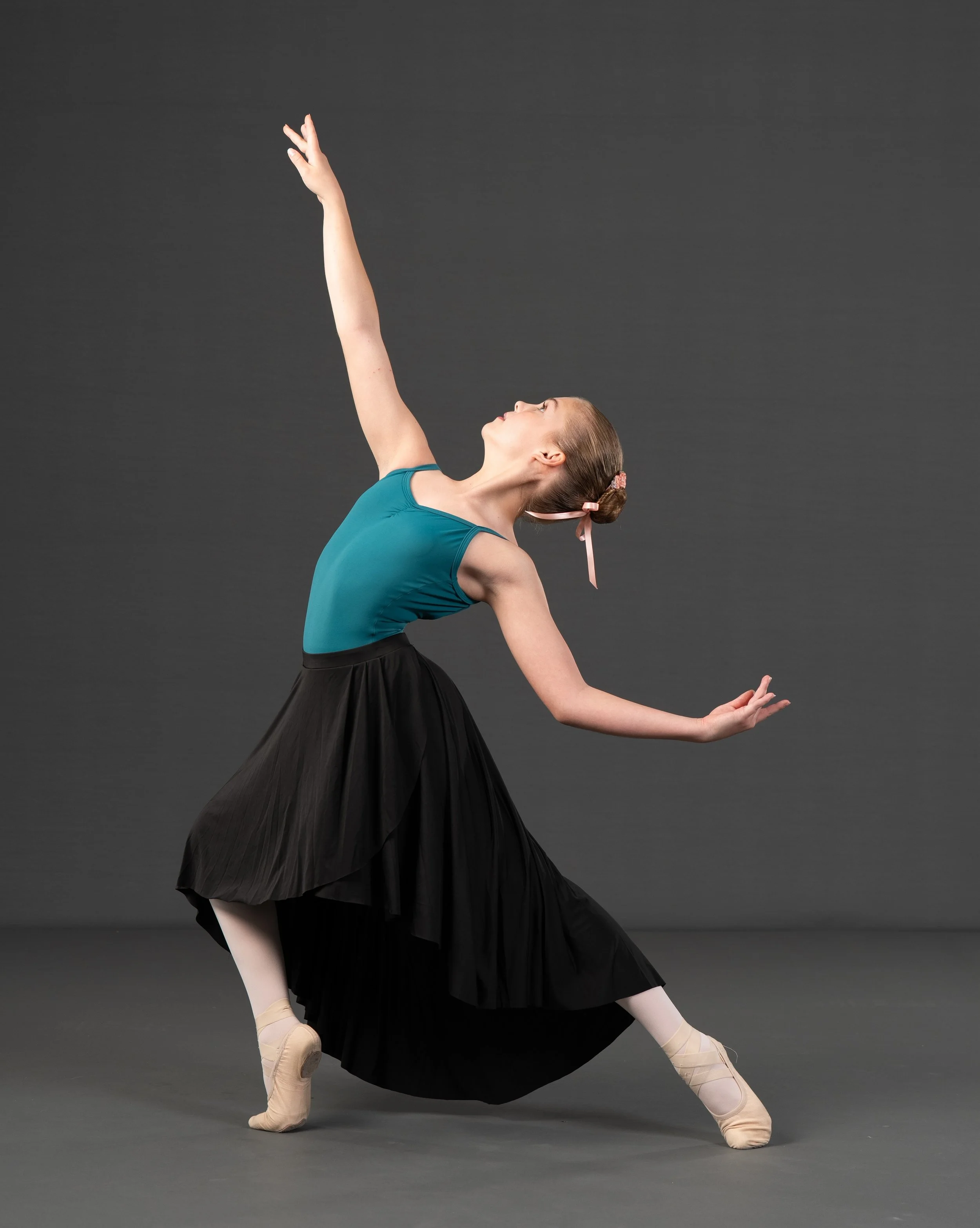 Ballet dancer in a pose with one arm raised and the other arm extended, wearing a teal leotard, black skirt, and satin ballet shoes, against a dark gray background.