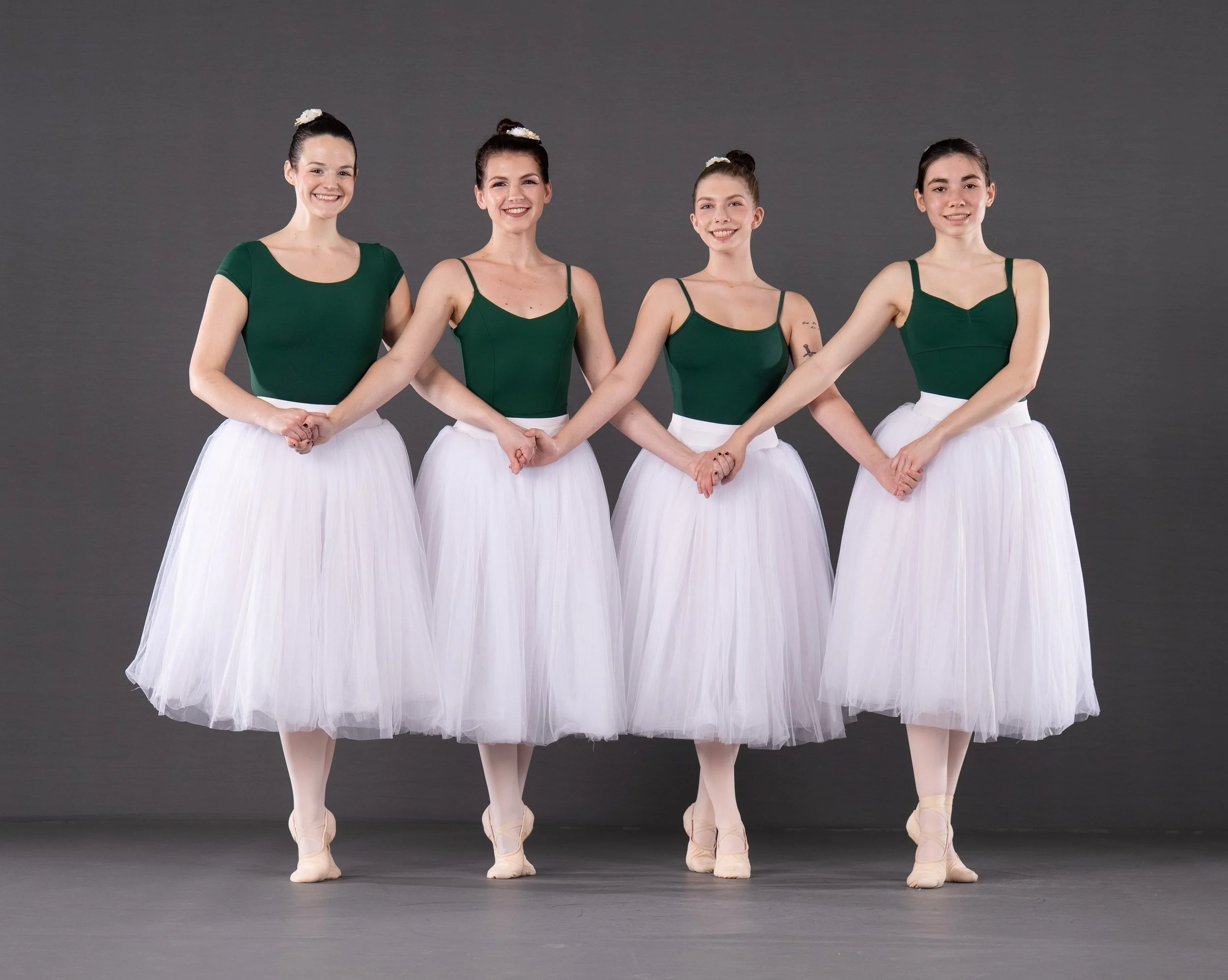 Four ballet dancers in green leotards and white tutus standing in a line holding hands against a gray background.
