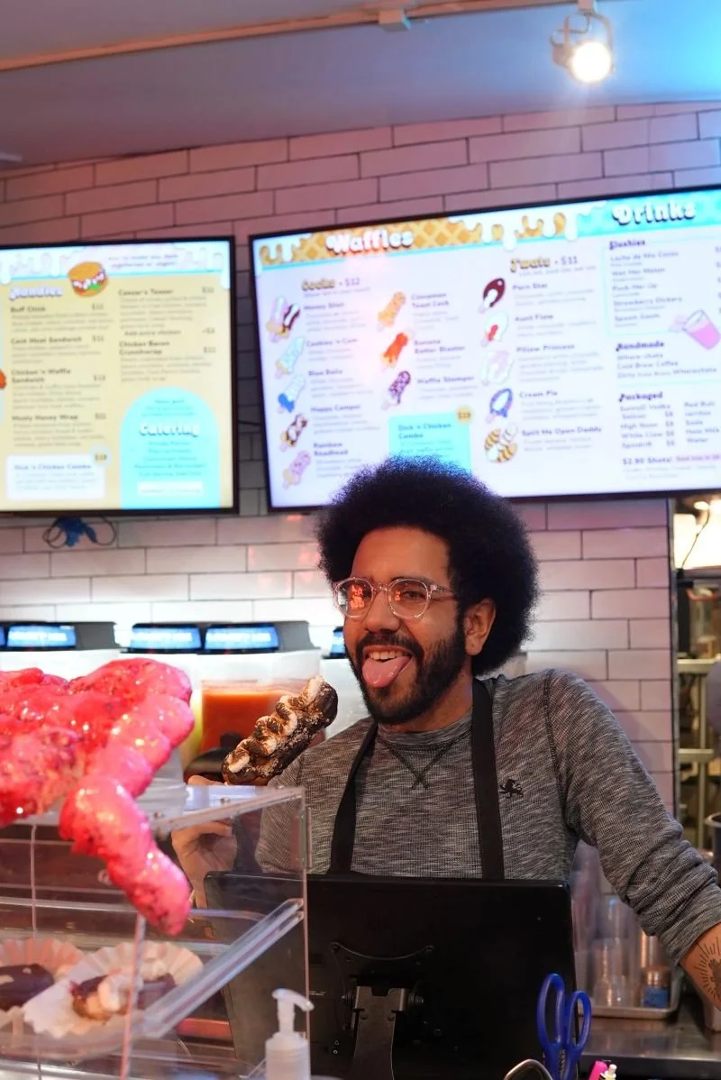 A man with glasses and an Afro hairstyle sticking out his tongue in front of a waffle shop. There are two menu screens in the background and waffle toppings in the foreground.