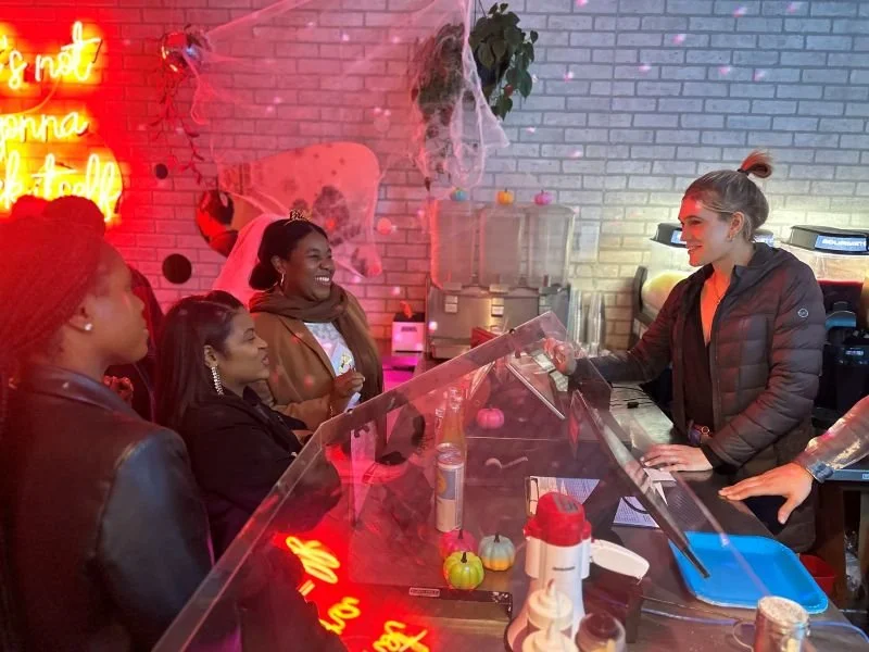 Four women are standing at a counter in a decorated cafe, with a bartender serving them. The decor includes a brick wall with Halloween decorations, string lights, and a neon sign. The women are smiling and engaging with the bartender.