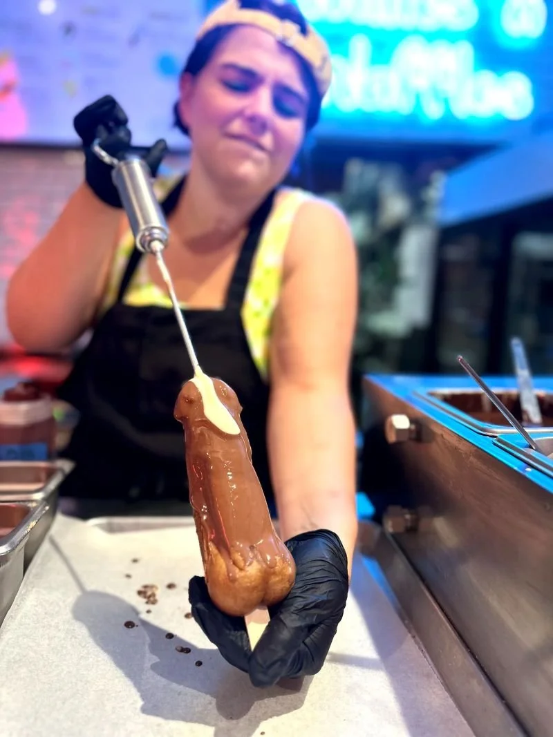 A woman wearing a yellow cap and black apron is holding a churro dipped in chocolate with a squeeze bottle of white sauce on it.