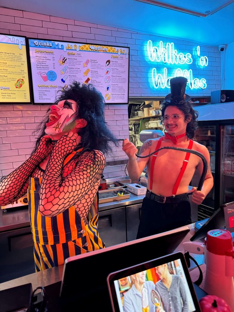 People dressed in circus-themed costumes at a donut shop, one with clown makeup and wig, the other wearing a top hat and suspenders, with a neon sign reading "Willes's Waffles" in the background.