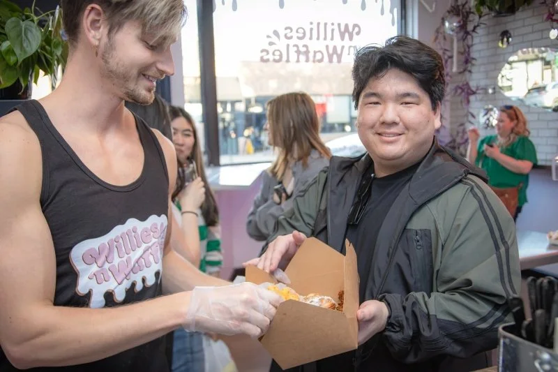 A smiling man receives a food order from a split hand wearing gloves inside a restaurant or cafe with large windows and several patrons in the background.