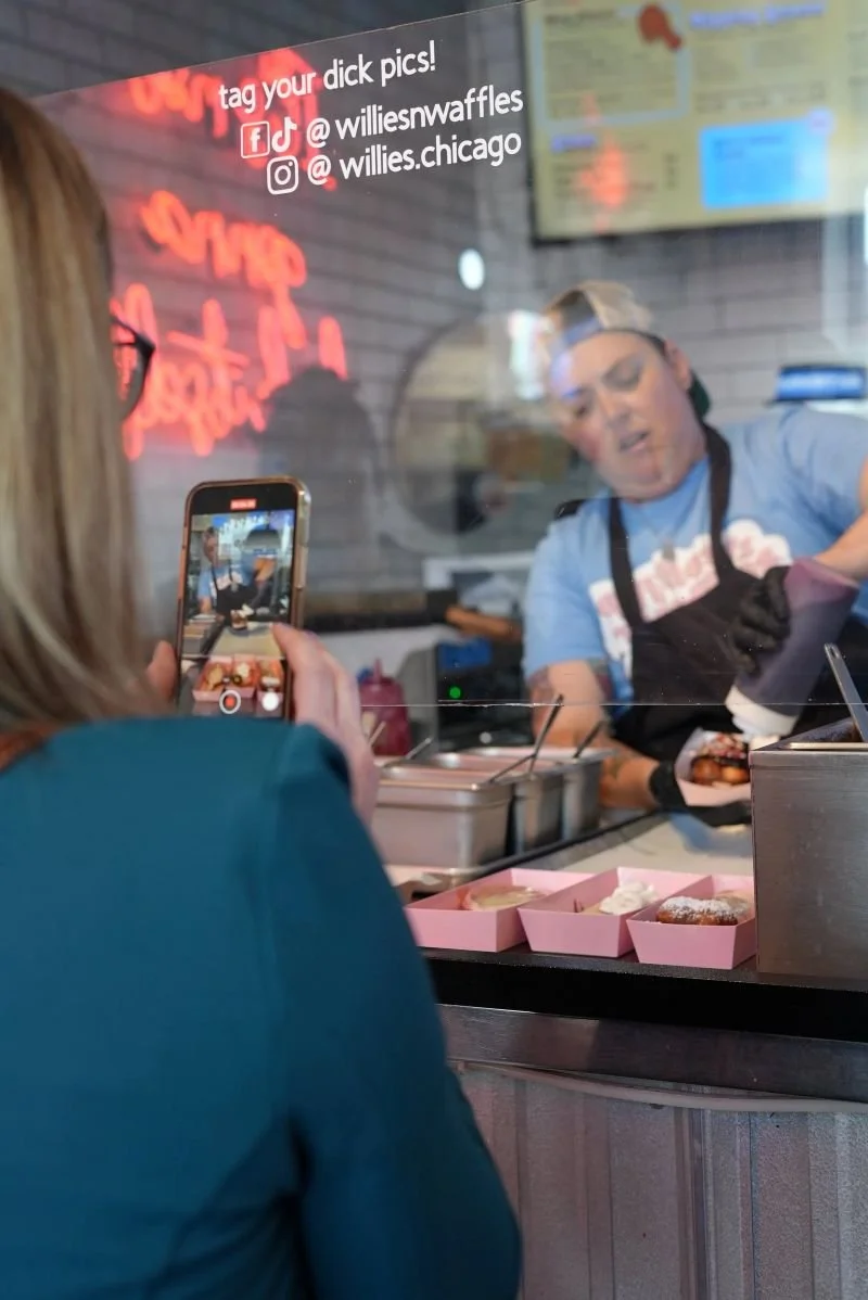 Person taking a photo of a worker preparing food at a counter in a restaurant. The worker is wearing a cap and apron, and there are boxes of food and a glass partition between them. Red neon signs and a digital menu are visible in the background.