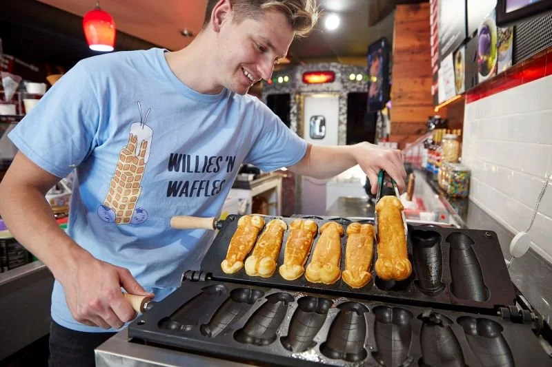 A young man cooking waffle ice cream cones on a waffle iron at an ice cream shop.