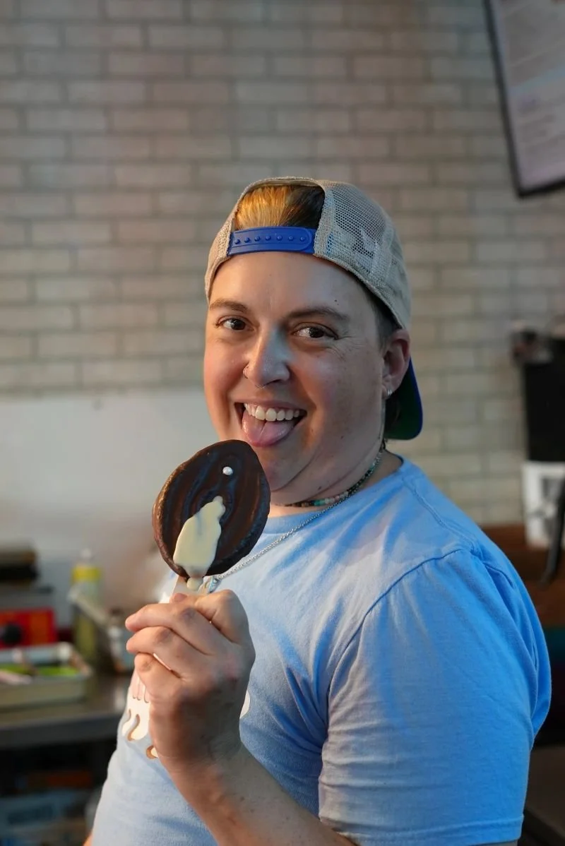 Young person smiling and sticking out tongue, holding a chocolate cookie with white filling, in an indoor setting with a brick wall background.