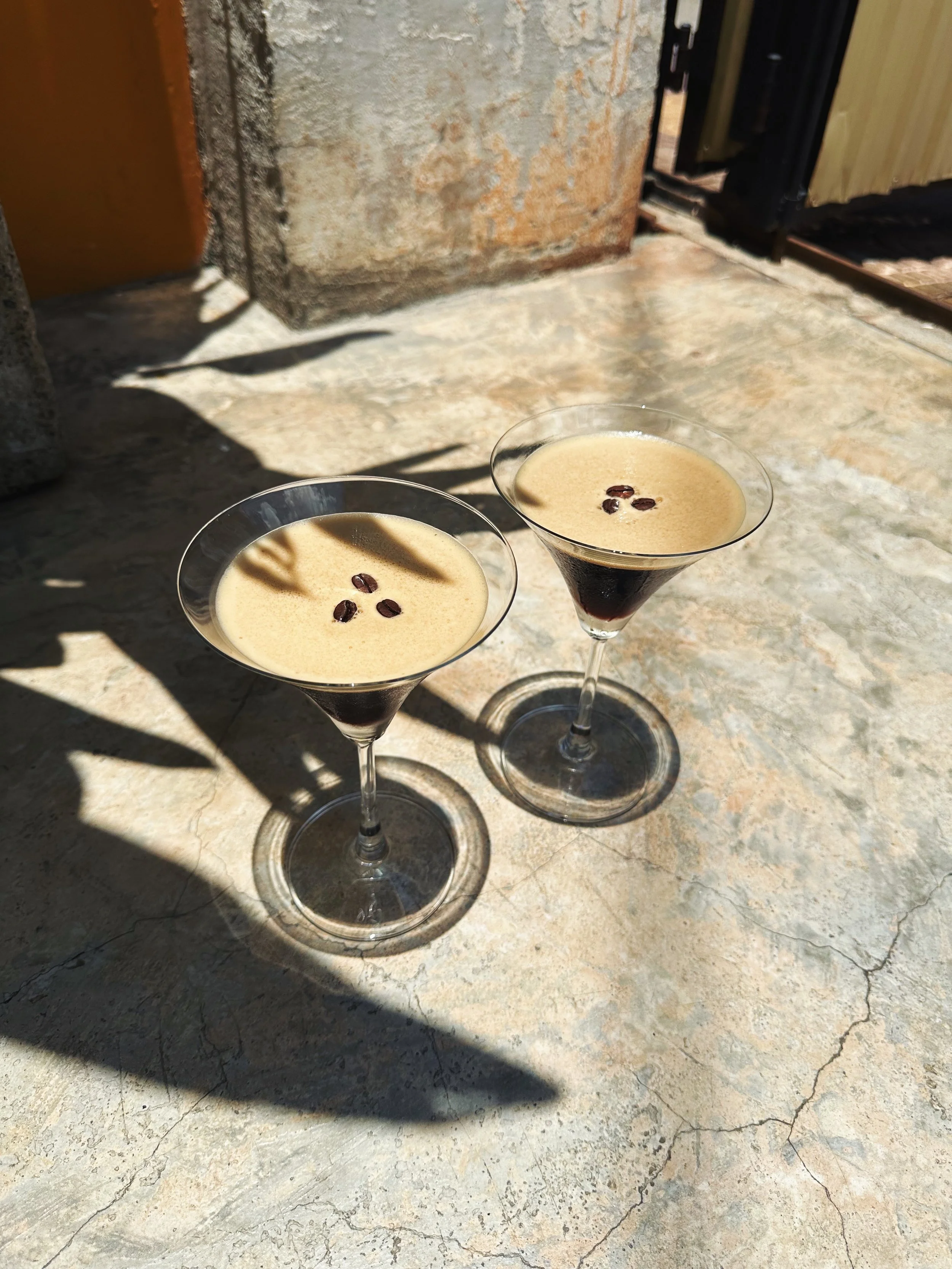 Two martini glasses filled with coffee and topped with coffee beans, placed outdoors on a stone surface with shadows.