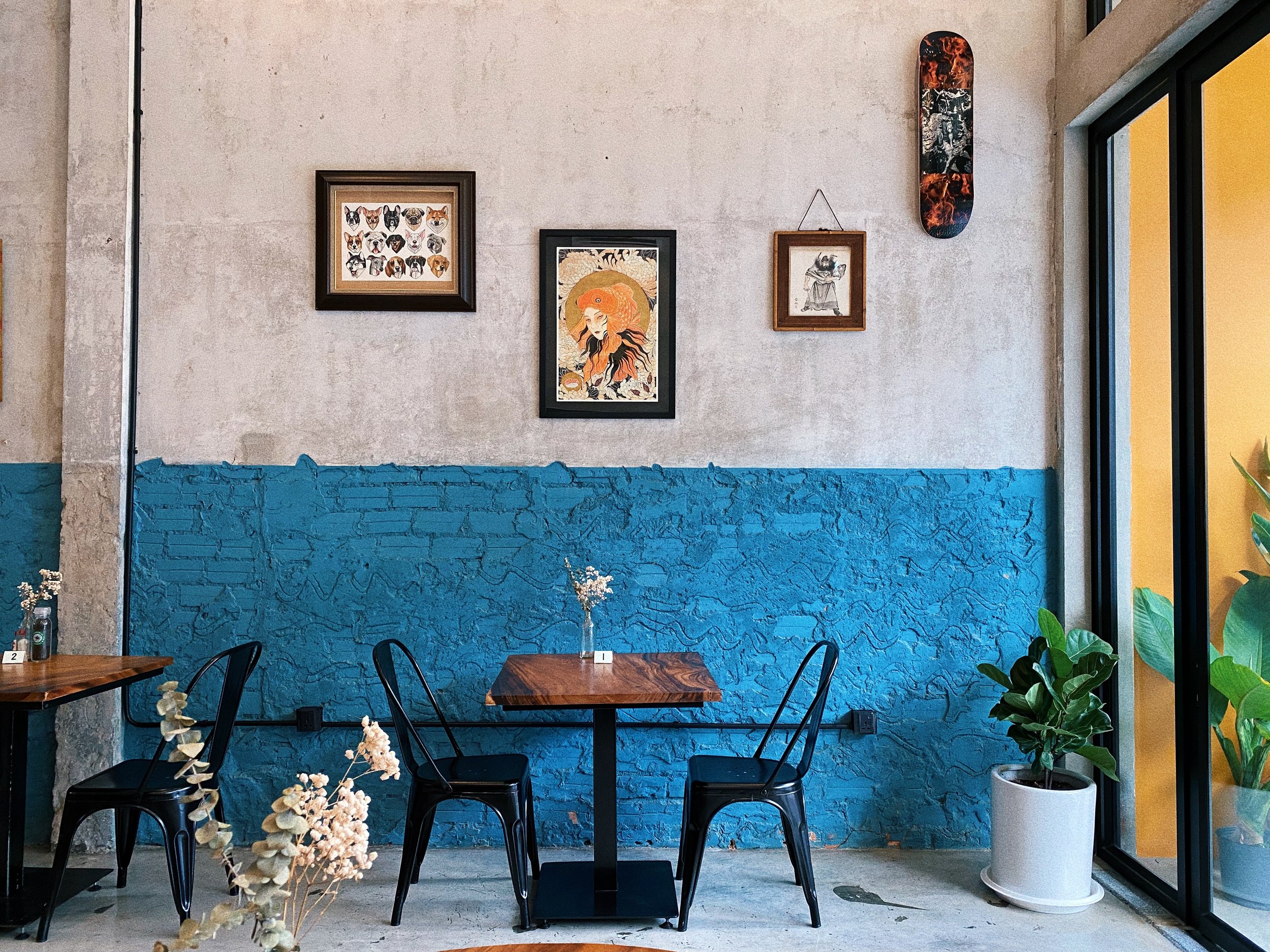 Interior of a modern café or restaurant with a concrete wall painted half green, decorated with framed artwork, a skateboard hanging, and a table with black chairs. There are plants on the right side of the image.