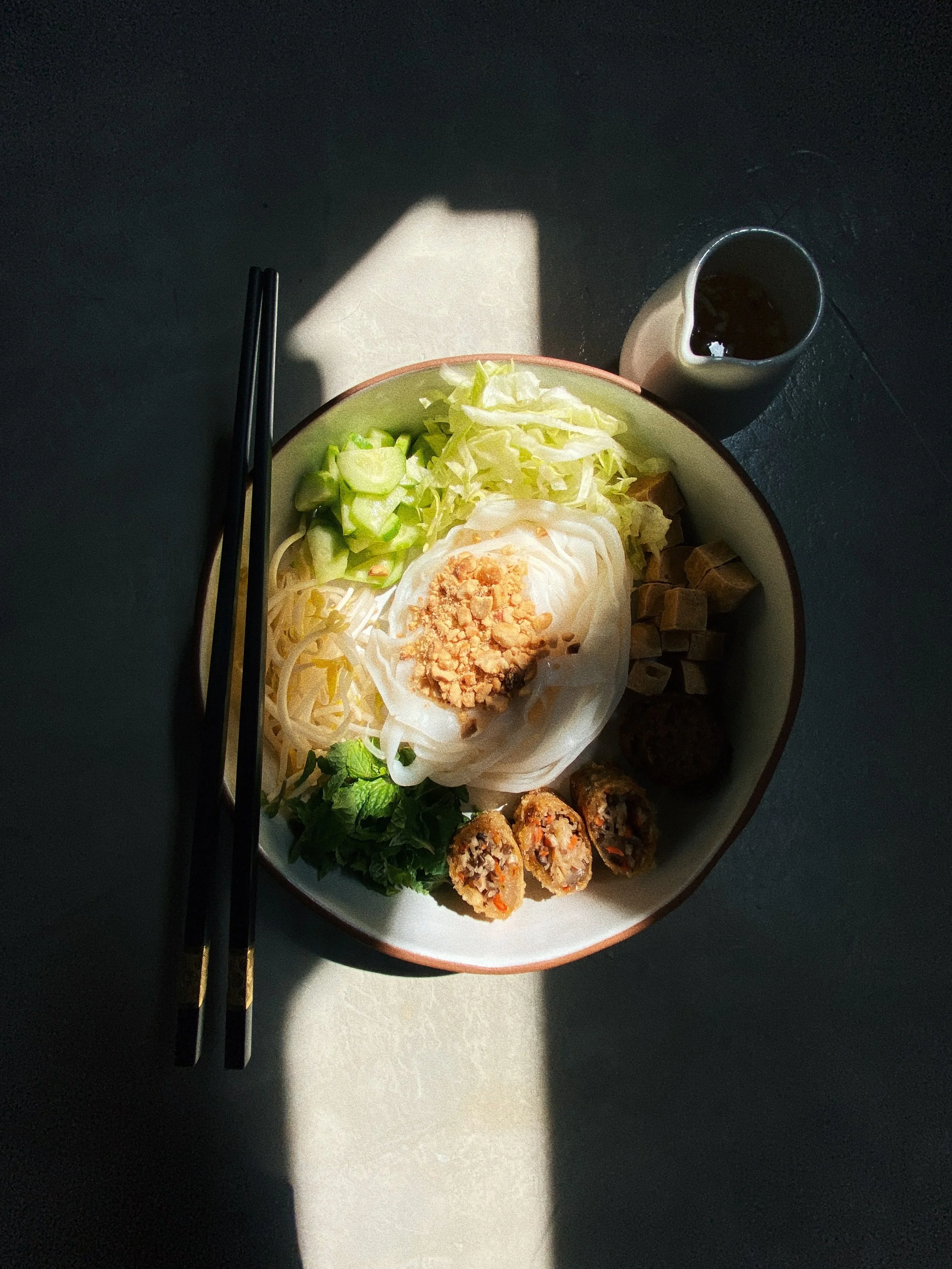 A bowl of Asian noodle soup with sliced cucumbers, shredded cabbage, a soft-boiled egg topped with crushed peanuts, and four pieces of cooked sushi rolls, accompanied by a small pitcher of dark soy sauce, chopsticks on the left side, set on a dark surface with a light reflection.