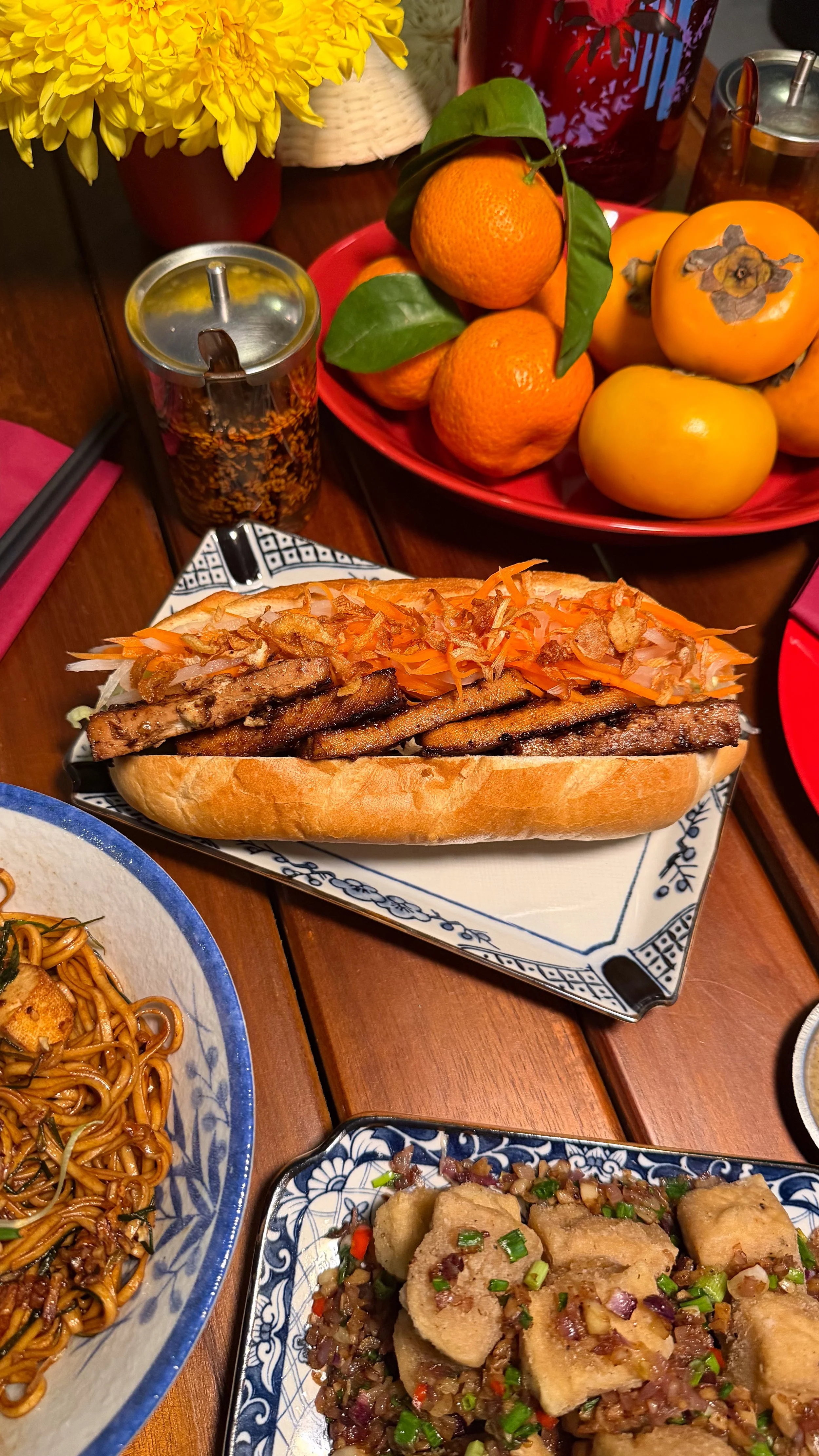 A hot dog with grilled meat, shredded carrots, and onions on a bun, surrounded by dishes of Asian cuisine, a bowl of stir-fried noodles, a vegetable dish with tofu, and a bowl of rice with vegetables. In the background, a bowl of mandarins, a vase wi