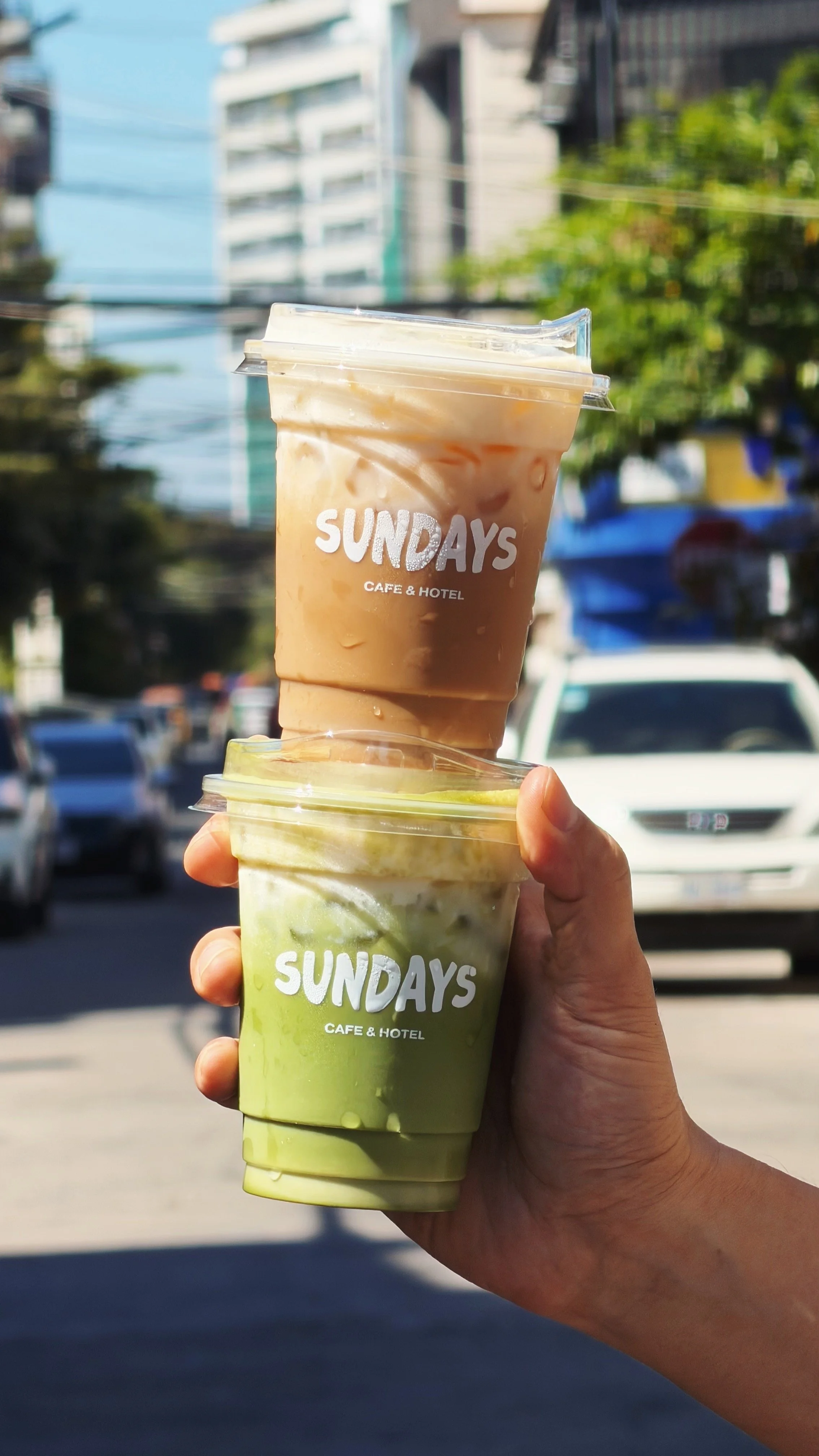 A person's hand holds three cups of beverages against an urban background with buildings and a street. The top drink is an iced coffee, the middle is an iced matcha latte, and the bottom is an iced green tea or matcha drink. All cups display the text 'SUNDAYS CAFE & HOTEL'.