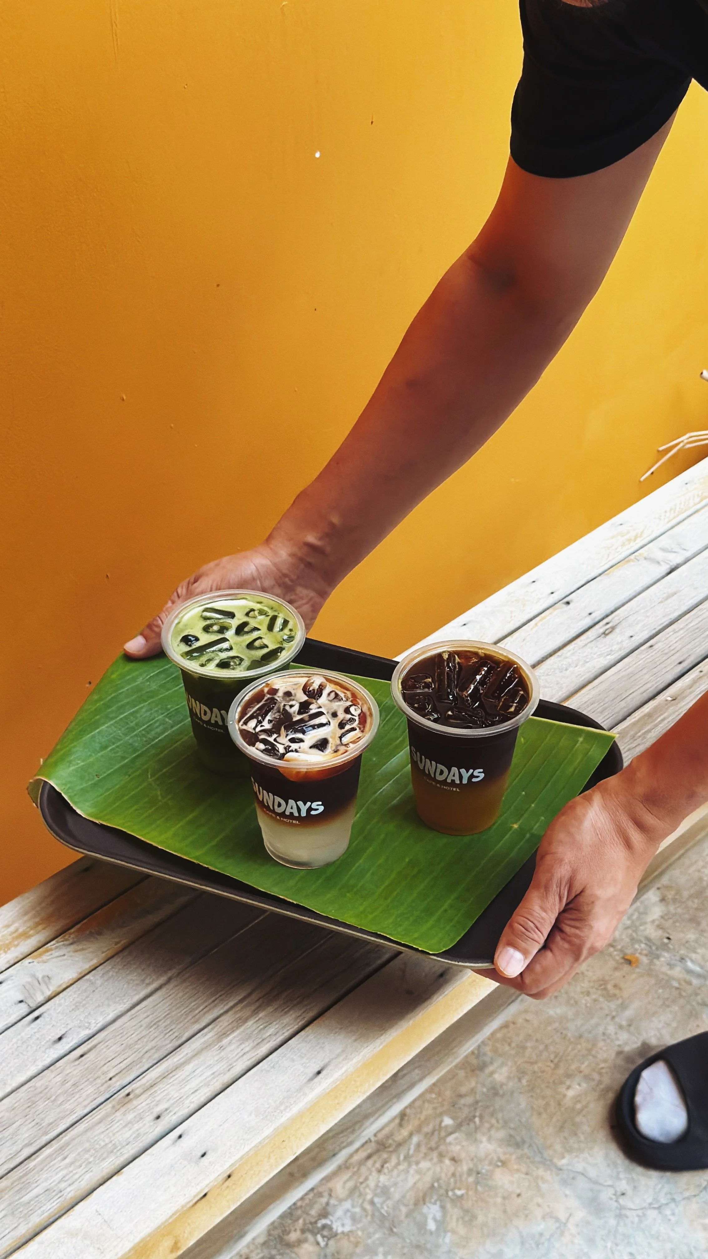 Person holding a tray with three iced drinks on a banana leaf, set on a wooden bench, against a yellow wall.