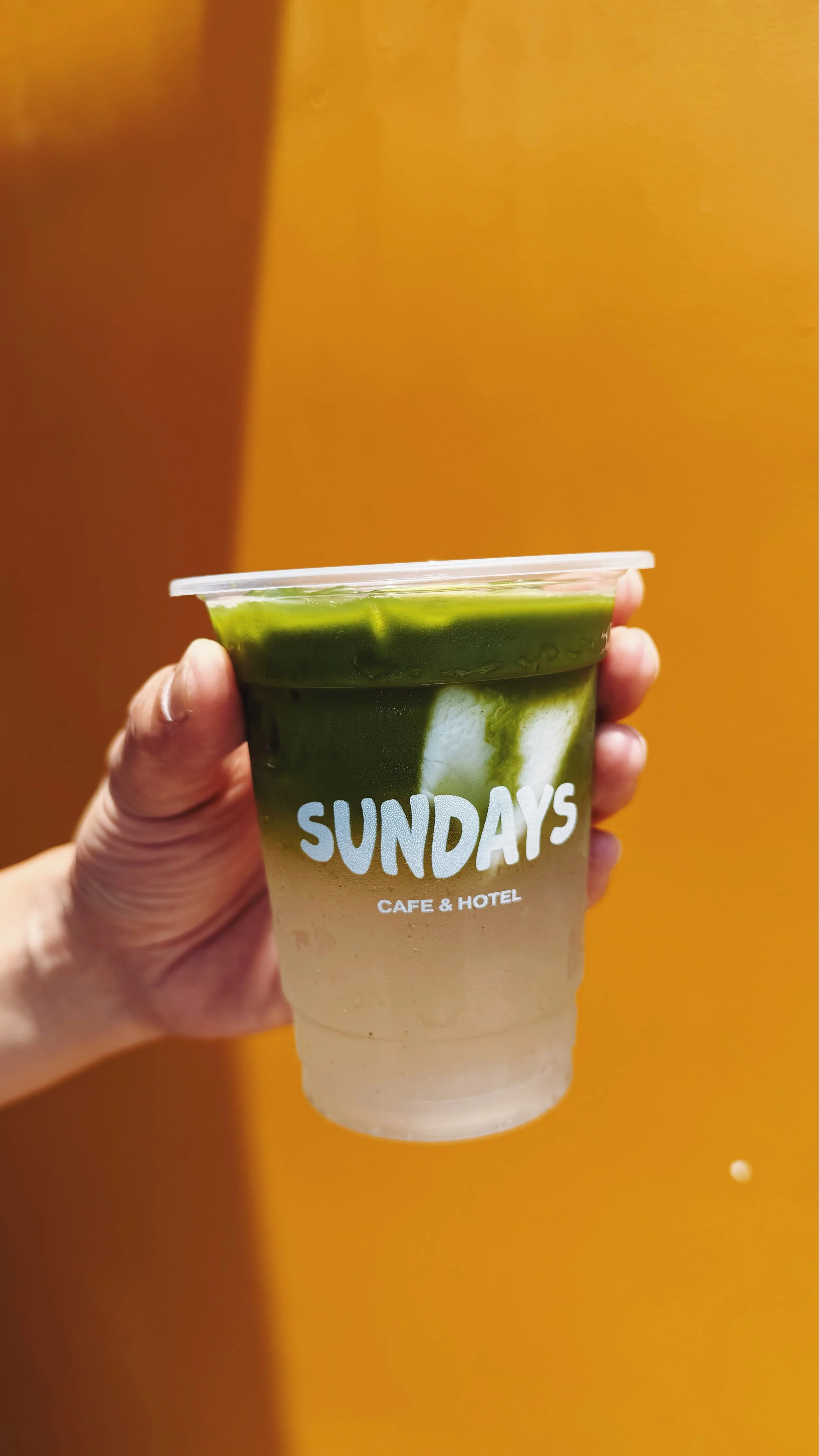 A hand holding a clear plastic cup with an iced matcha coconut water drink, against a sunny yellow wall. The cup has the words 'sundays cafe & hotel' printed on it in white
