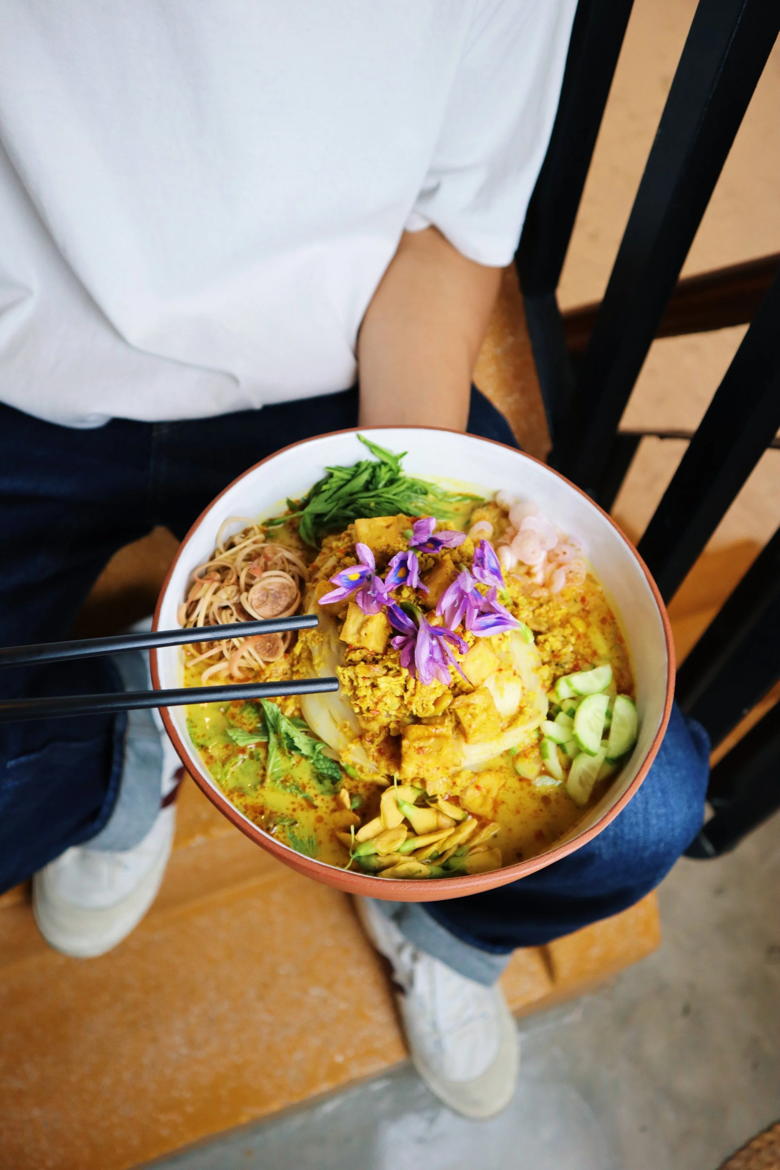 A person sitting on a yellow terrazzo staircase holding a large bowl of a colorful Cambodian dish called Nom Banh Chok Samlor Namya, which consists of a red curry with vermicelli noodles, fresh cucumber, bean sprouts & lotus stems