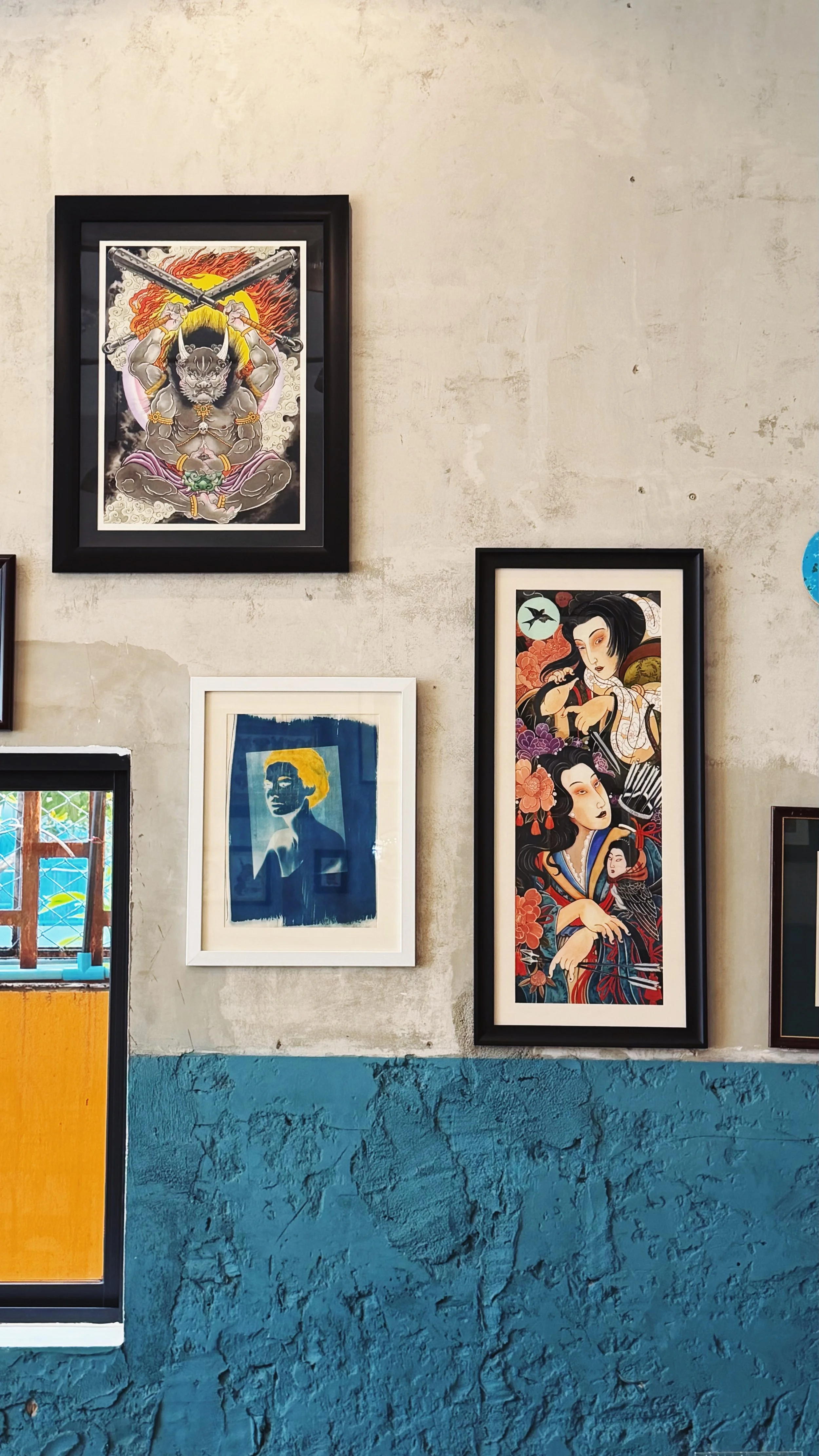 Wall decor with four framed artworks, including a traditional Japanese style warrior with crossed weapons, a blue-toned silhouette of a woman, and two artworks featuring female figures with floral and nature motifs, against a textured beige and blue wall.