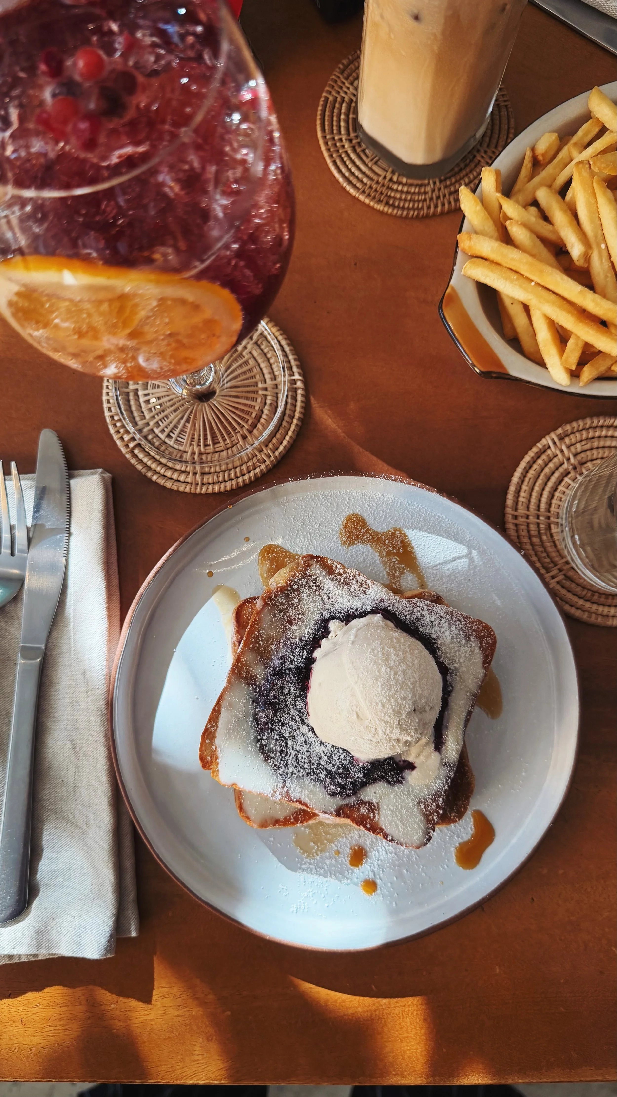 Plate with Belgian waffle topped with whipped cream, icing sugar, and syrup, accompanied by a glass of iced coffee, a glass of berry soda, and a basket of French fries.