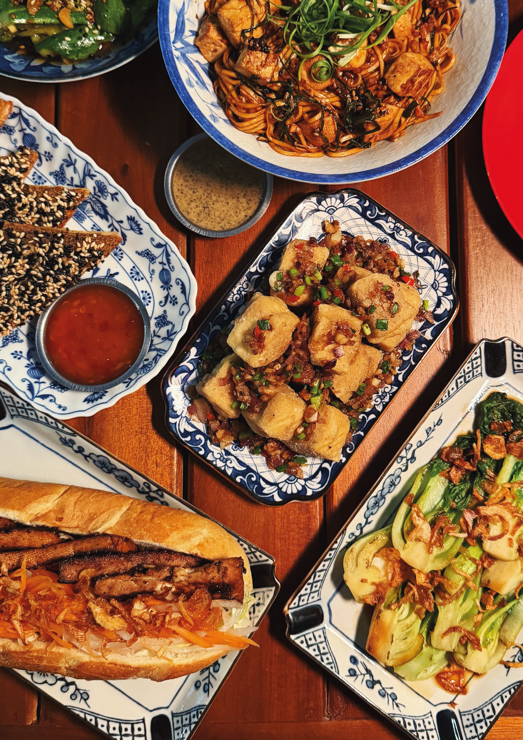 A variety of Asian dishes on decorative plates, including fried tofu with sauce, noodle stir-fry with vegetables and tofu, spring rolls with dipping sauce, and a sandwich with shredded vegetables.
