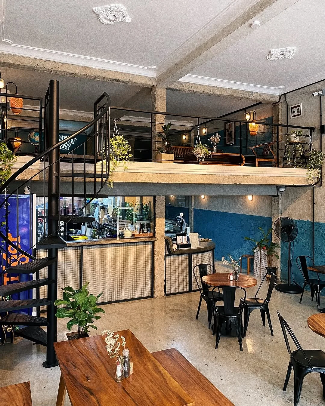 Interior of a modern cafe with wooden tables, black chairs, potted plants, a staircase, a mezzanine level with additional seating, string lights, and a counter area in the background.