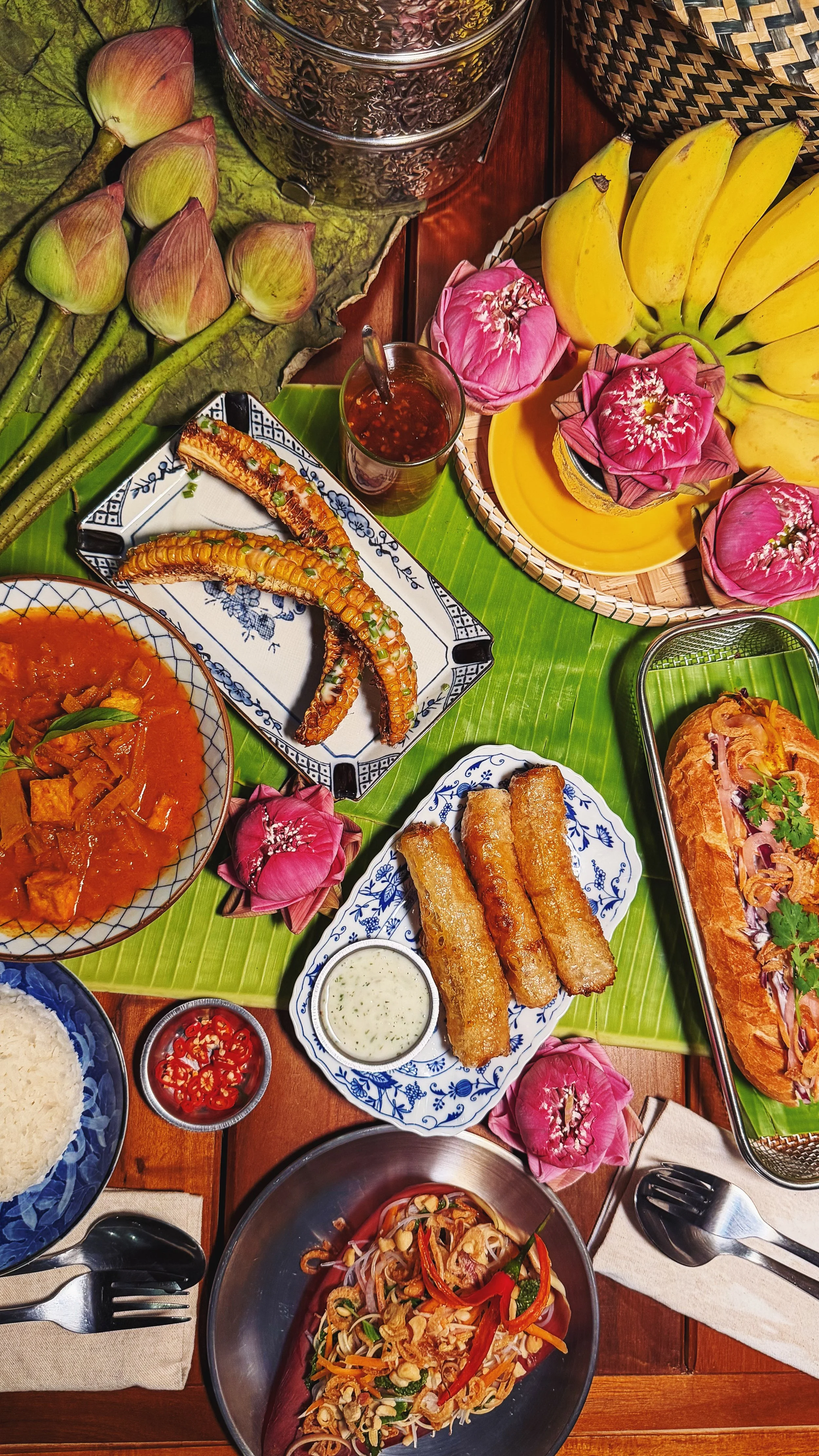 A variety of creative Cambodian food dishes including spring rolls, a vegetable curry and corn ribs, arranged on a table with utensils, bowls, sauces, and decorative lotus flowers and banana bunches & woven baskets.