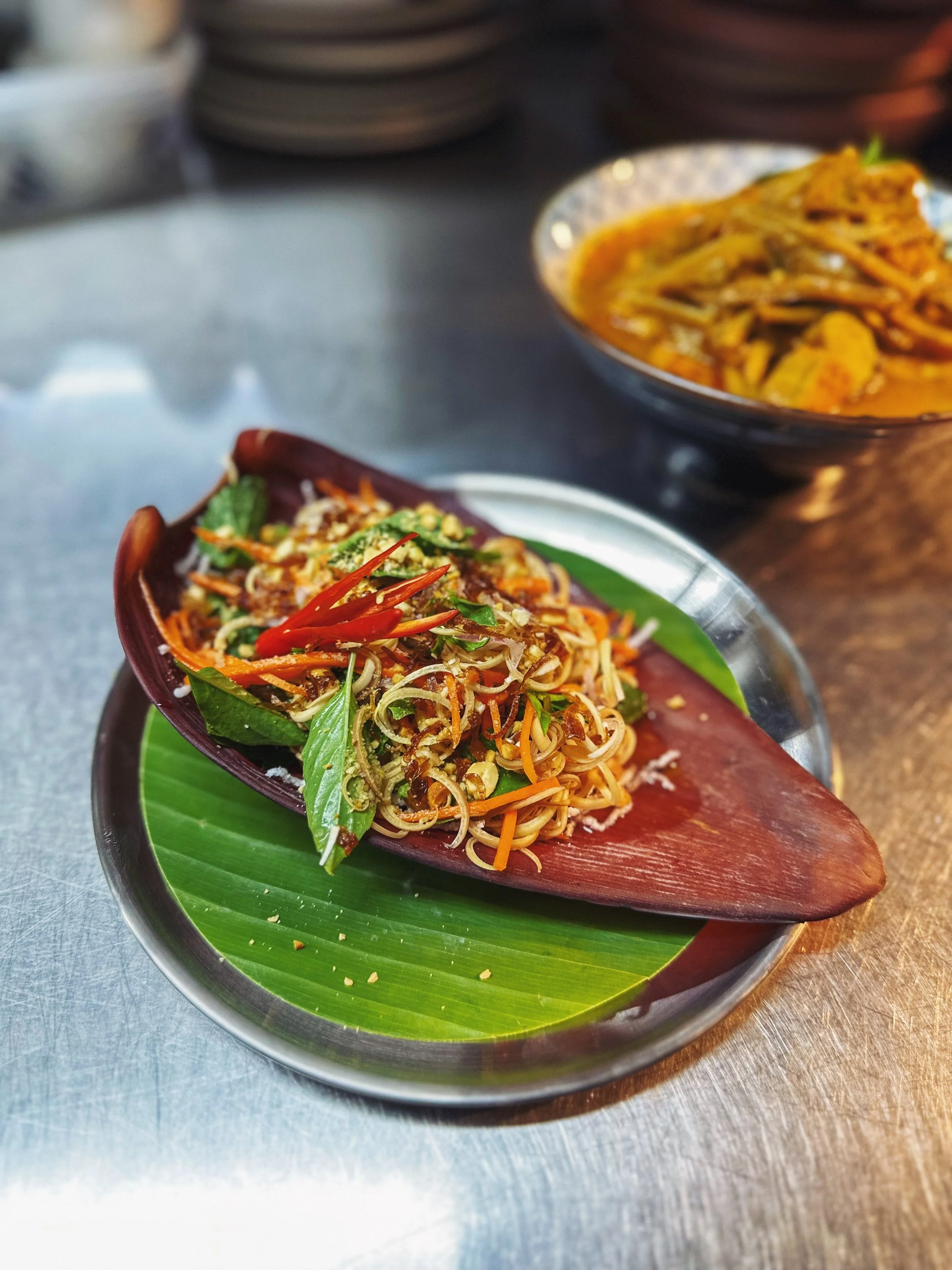 Close-up of a serving of spicy stir-fried noodles with vegetables and herbs on a banana leaf, placed on a metal tray, with another dish of curry in the background.