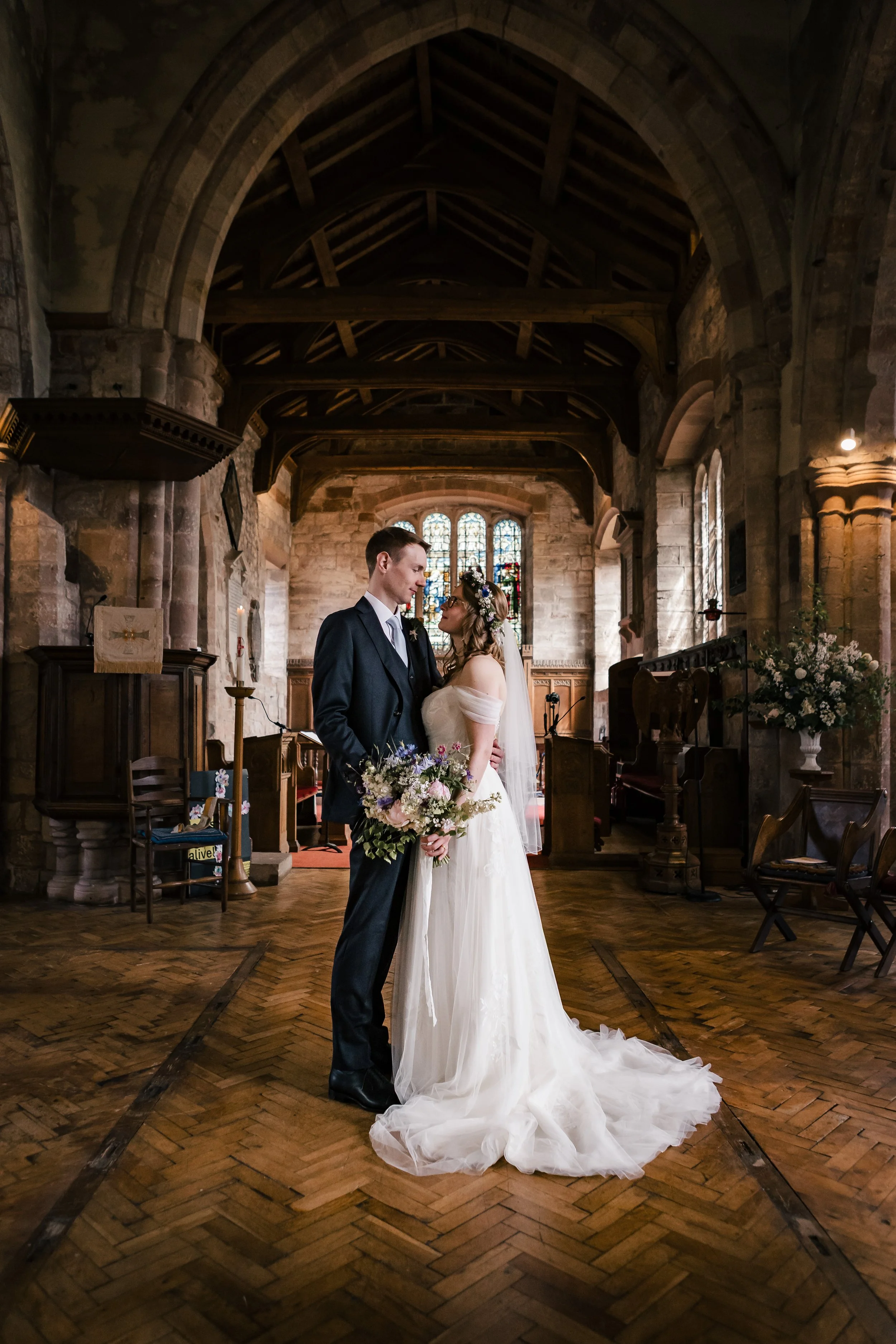 A bride and groom standing close inside a historic stone church with wooden beams, stained glass windows, and wooden pews and chairs. The bride is holding a bouquet and wearing a white wedding gown with an off-the-shoulder design and a floral crown.
