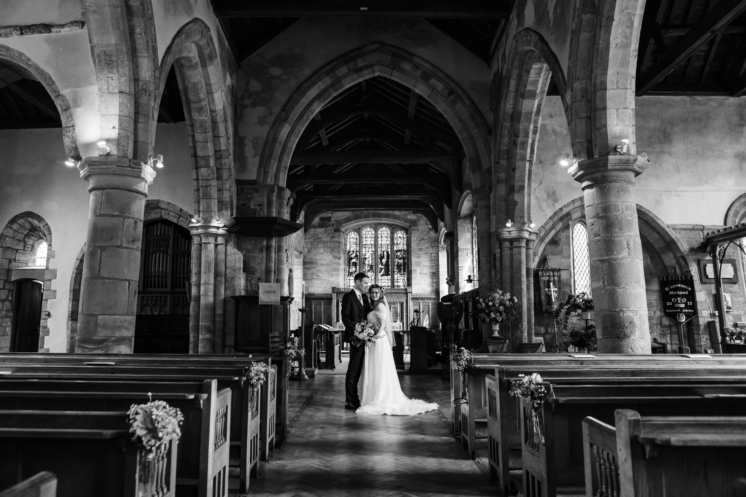 A black and white photo of a bride and groom standing in the center aisle of a historic church, with stained glass windows in the background and decorated pews along the aisle.