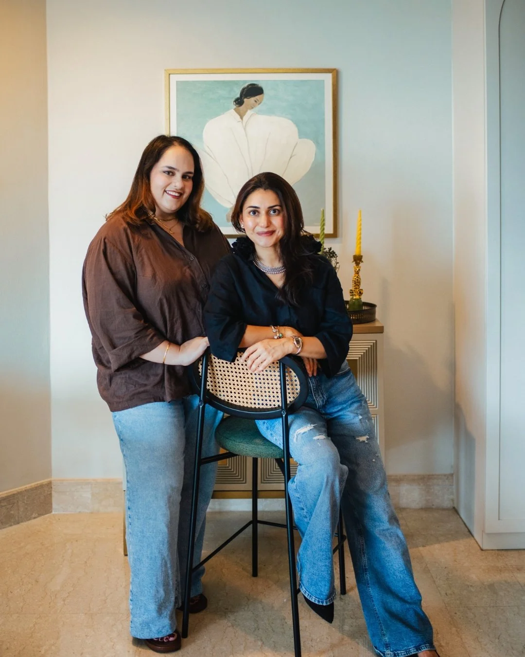 Two women smiling and posing for a photo indoors, one is standing, and the other is sitting on a chair with a green cushion. Behind them is a framed artwork of a woman with a large white flower, and a small cabinet with yellow candles and decorative items.