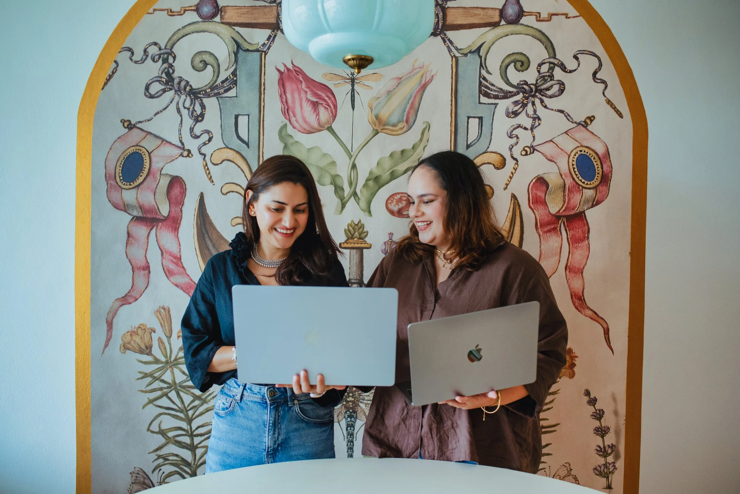 Two women standing together, smiling and looking at a laptop. They are in front of a painted mural with flowers and botanical illustrations.