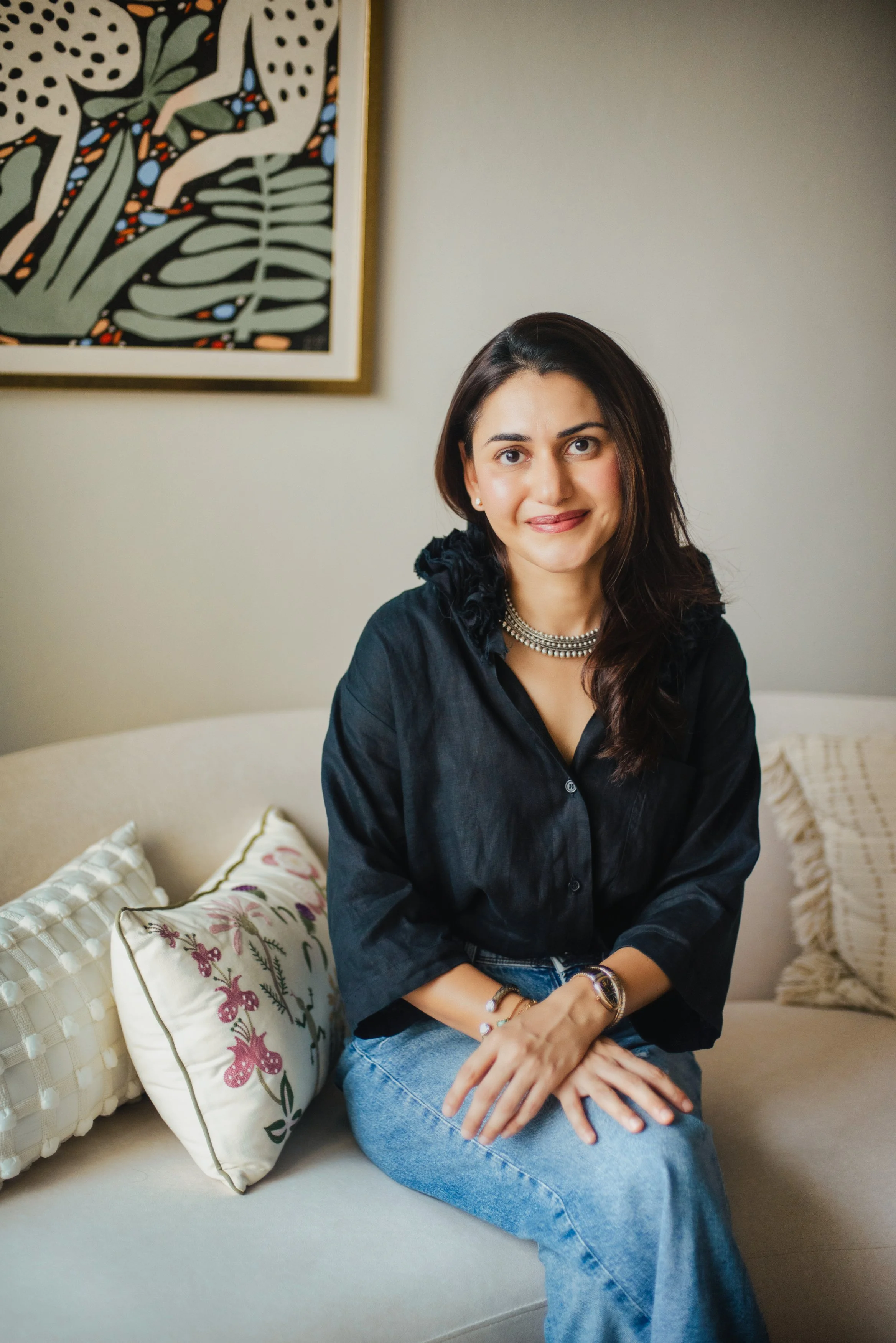 A woman sitting on a beige sofa, wearing a black blouse and jeans, surrounded by decorative pillows and in front of a colorful abstract artwork on the wall.