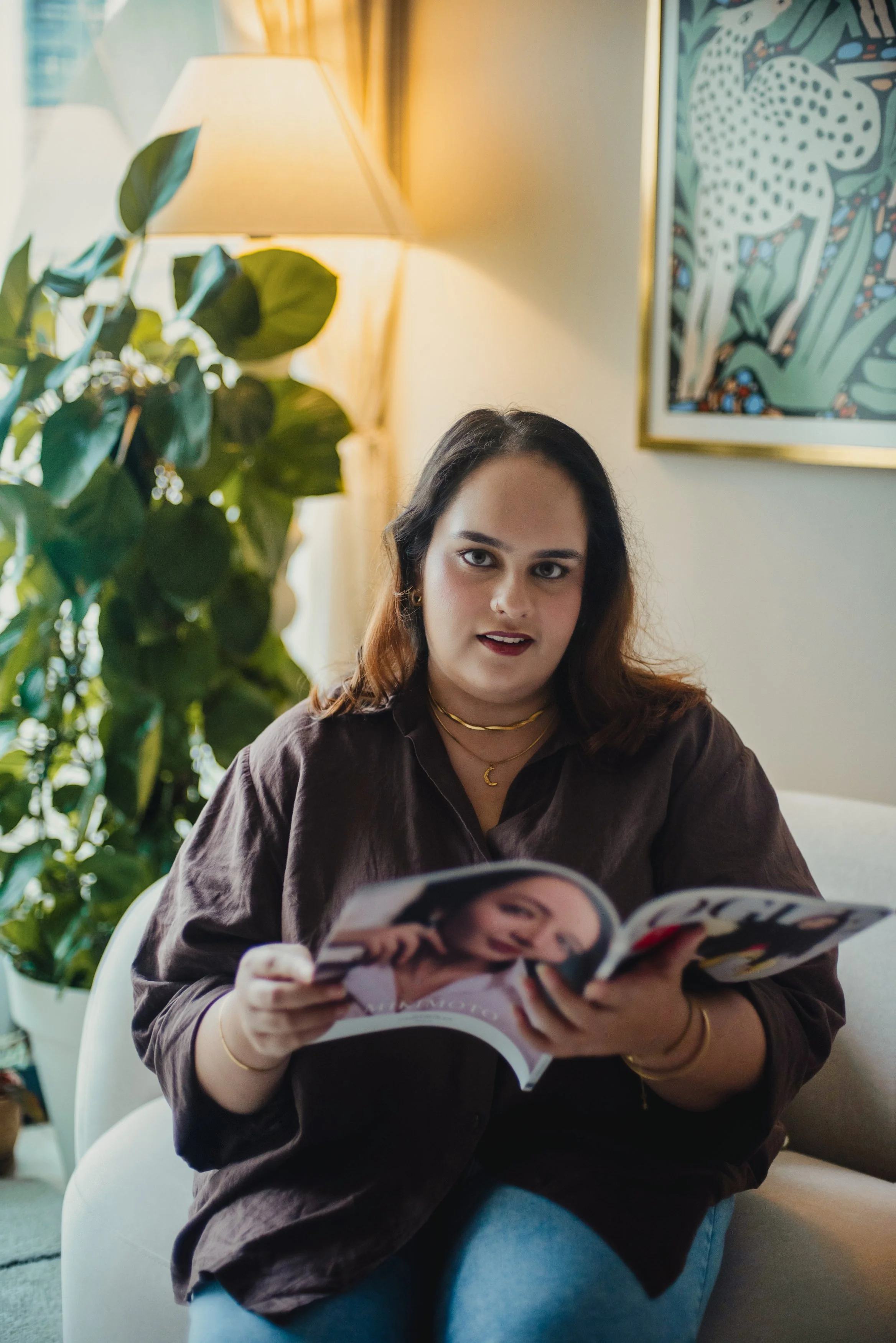 A woman with dark hair, wearing a brown shirt, gold jewelry, and red lipstick, sitting in a well-lit room reading a magazine. Behind her are a large leafy green plant, a framed colorful abstract art piece, and a warm-colored table lamp.