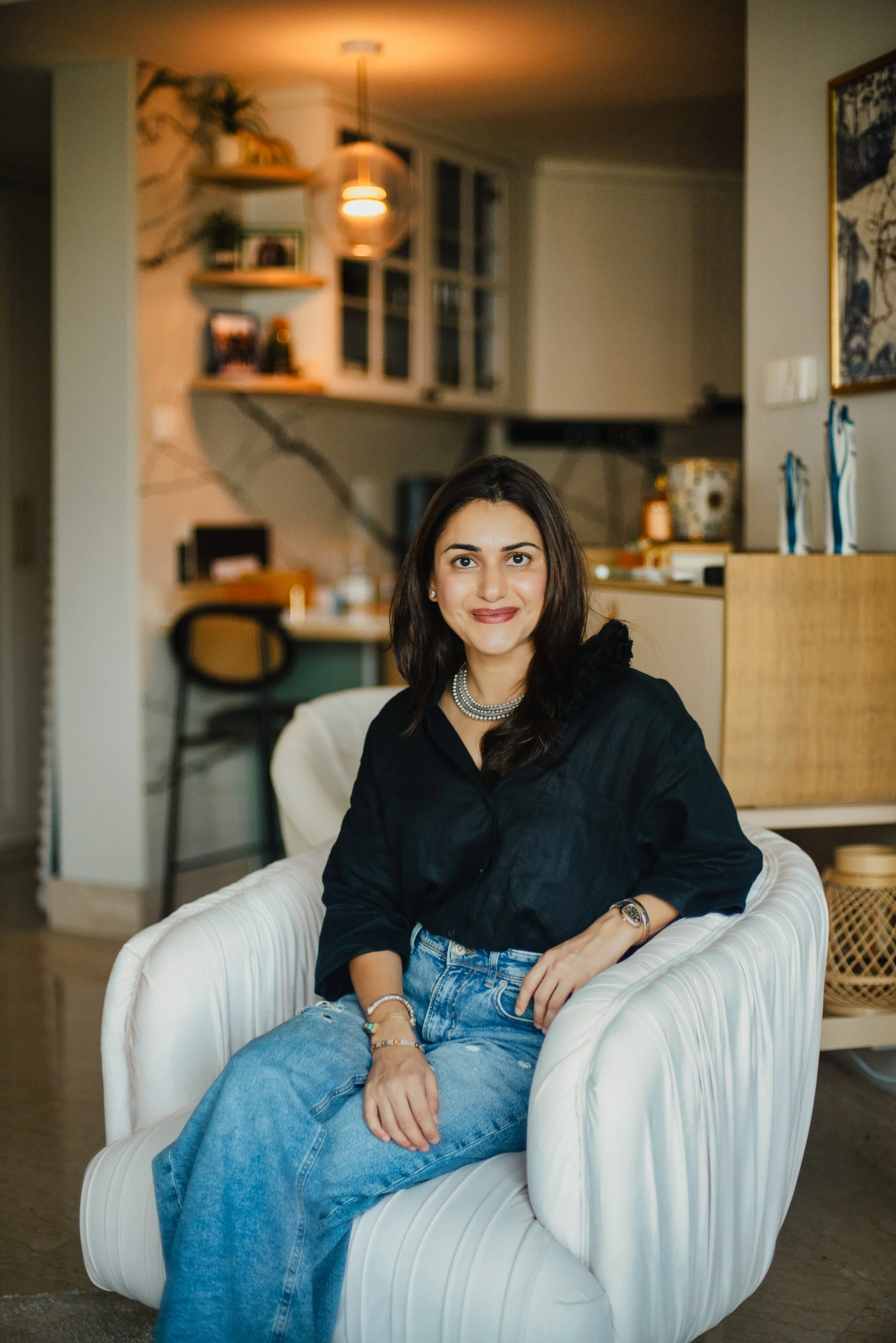 A woman with dark brown hair, wearing a black top, light blue jeans, and accessories, sitting comfortably in a white armchair in a warmly lit living room with decorative items and shelves in the background.