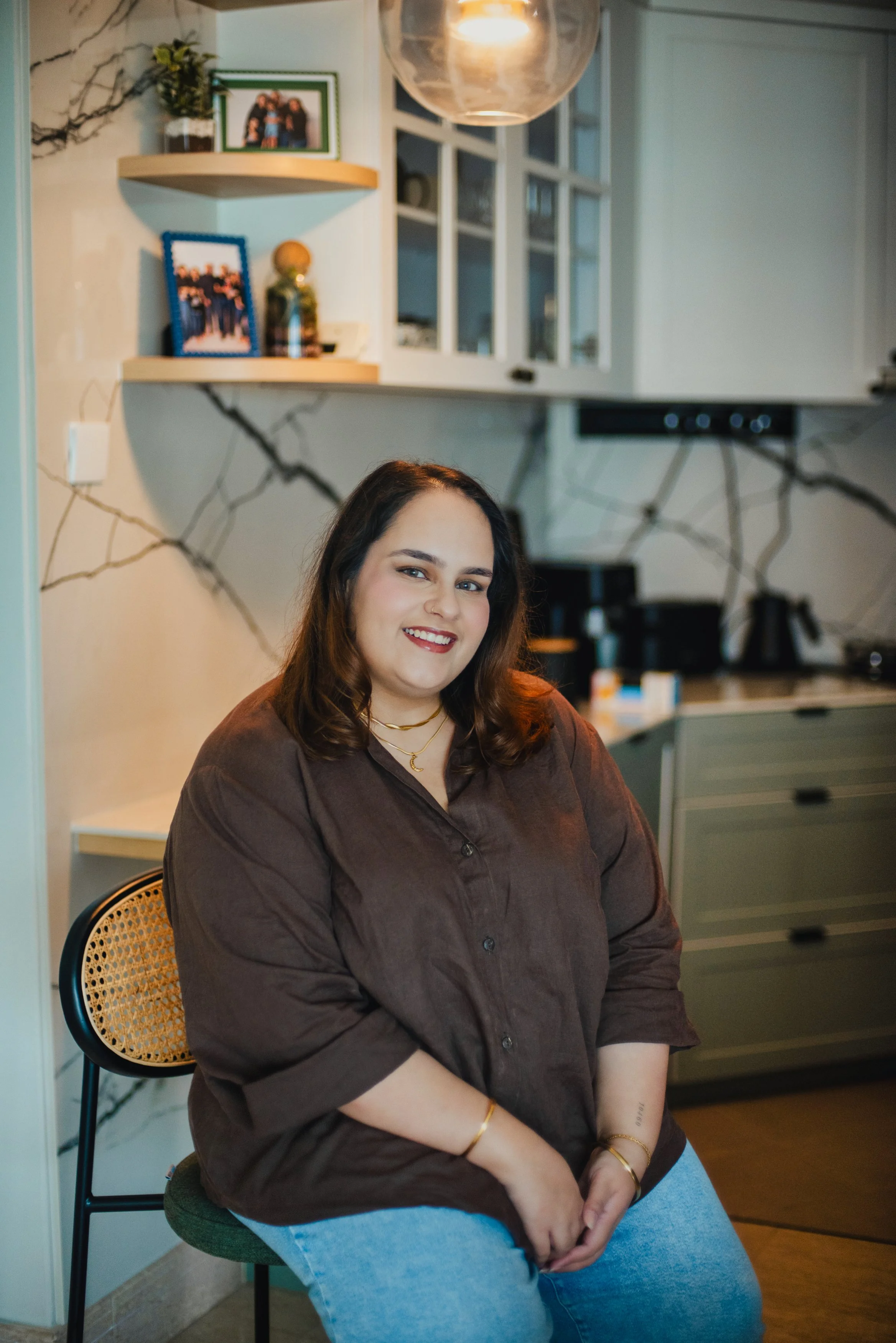 A woman with shoulder-length brown hair and a smile, wearing a brown button-up shirt and light blue jeans, sitting on a green chair in a kitchen with a marble-patterned wall and shelves with framed photographs and decorative items.