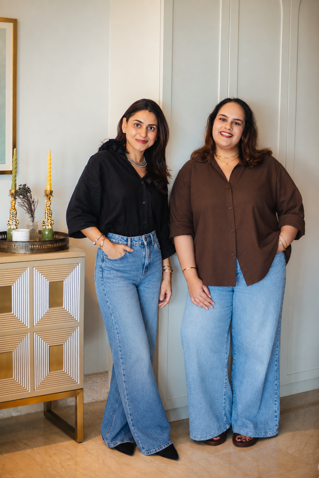 Two women standing indoors, smiling, dressed in casual outfits with wide-legged jeans and button-up shirts, posing near a decorative side table with candles and decorative items.
