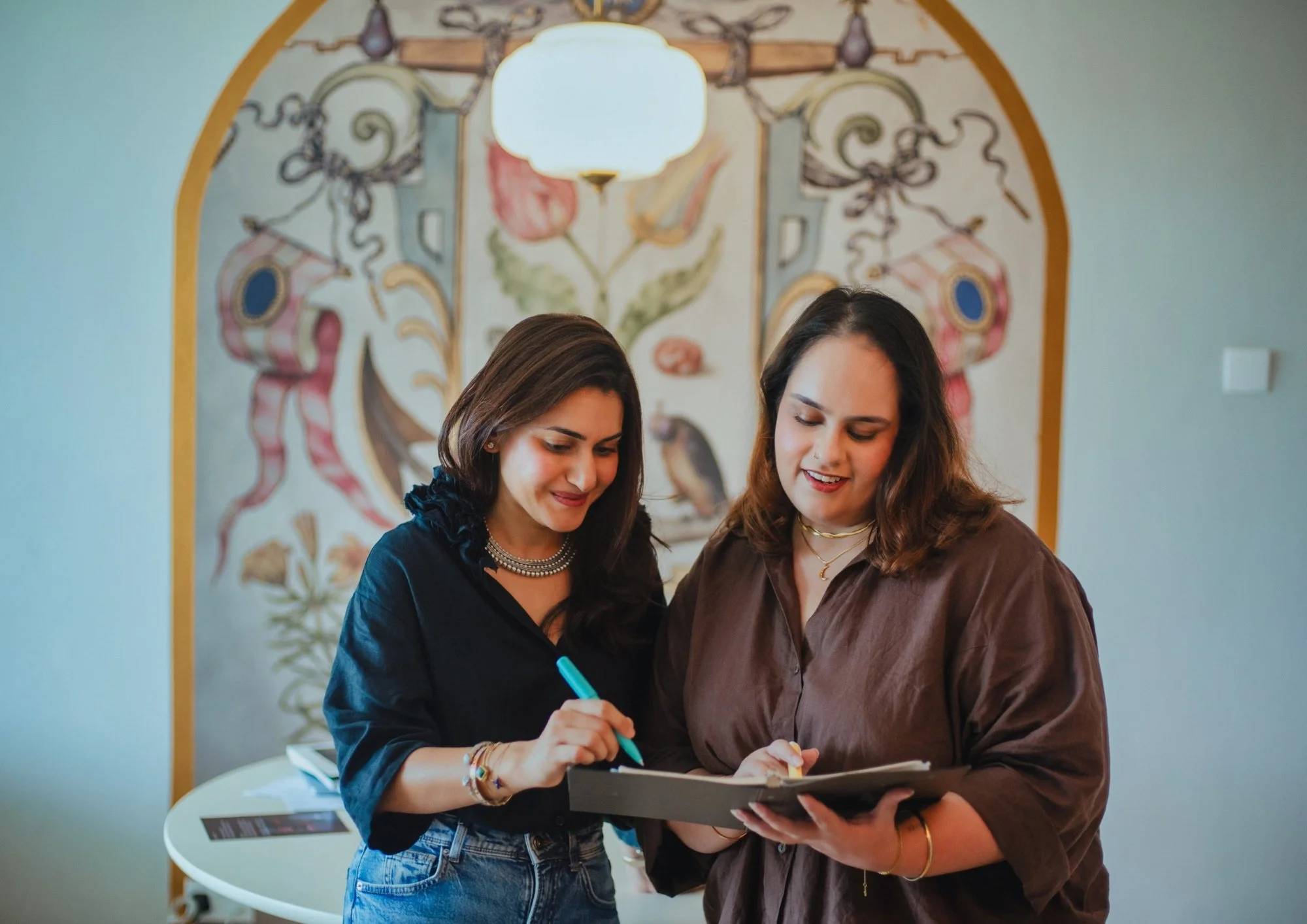 Two women standing at a table, signing a document together in an indoor setting with a decorative floral mural on the wall behind them.