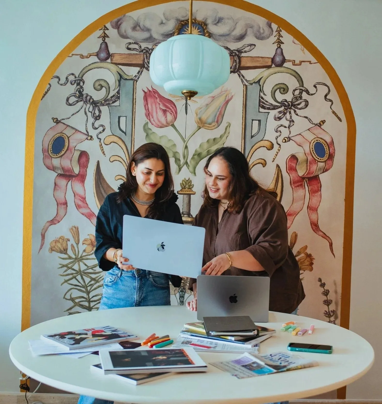 Two women standing behind a round white table, looking at a laptop and discussing. The table has magazines, papers, chargers, and colorful chalks. Behind them is a decorative mural with floral and ribbon designs, a cloud and sun in the center, and a hanging light fixture.