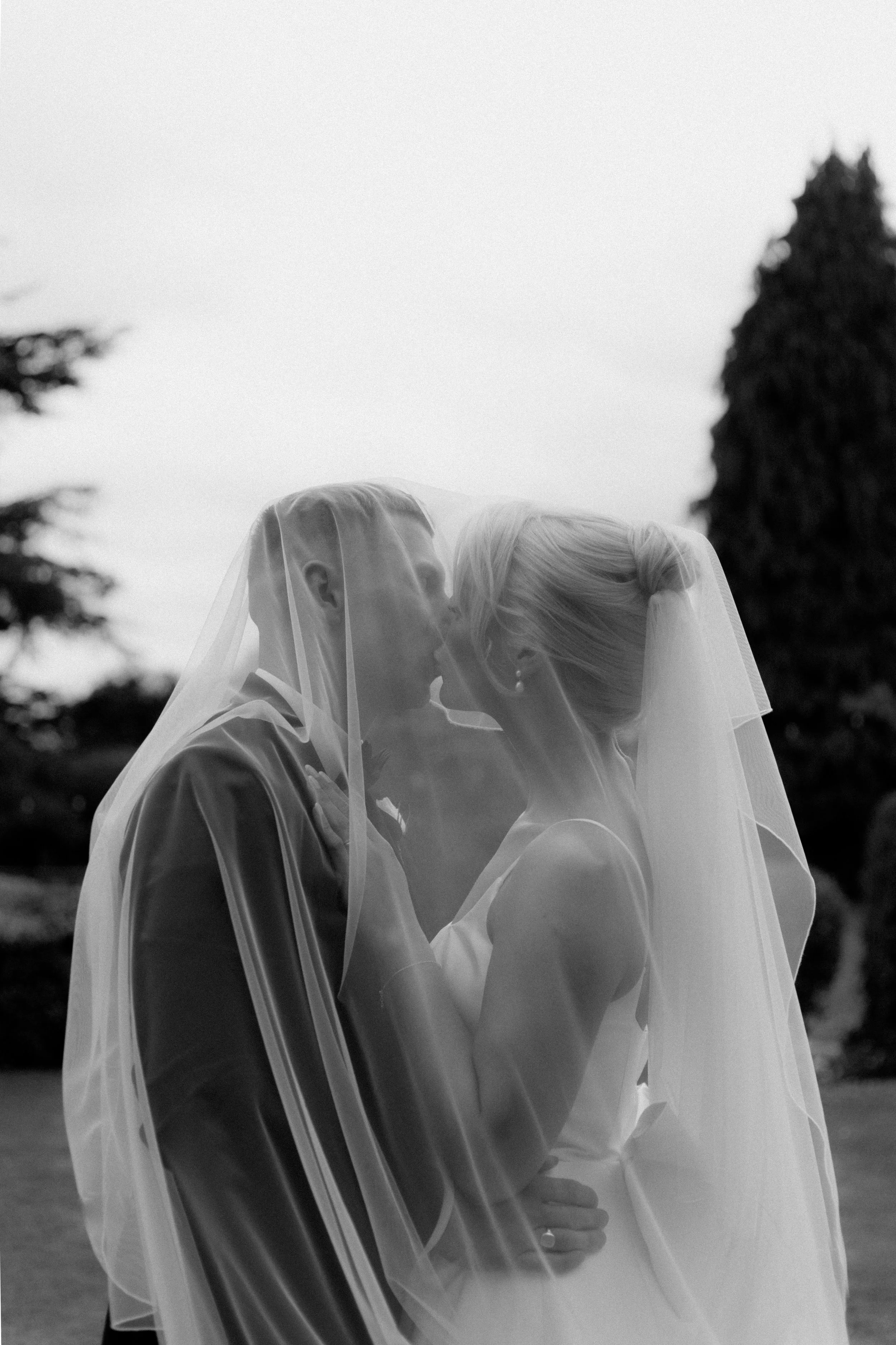 Black and white photo of a bride and groom holding each other under a veil outdoors.