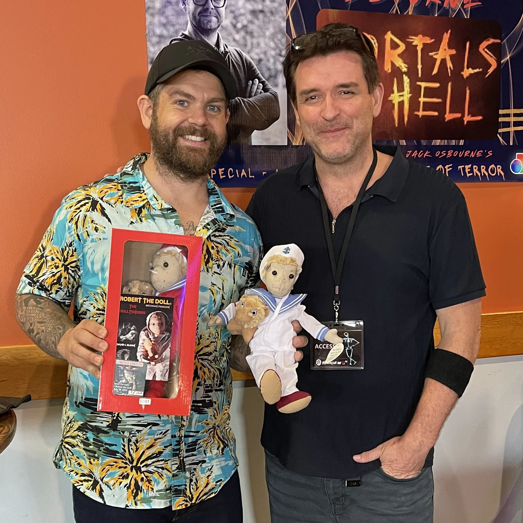Paranormal experts David L. Sloan and Jack Osbourne holding plush Robert the Doll toys. Behind them are posters, including one for 'Horrors of Hell.'
