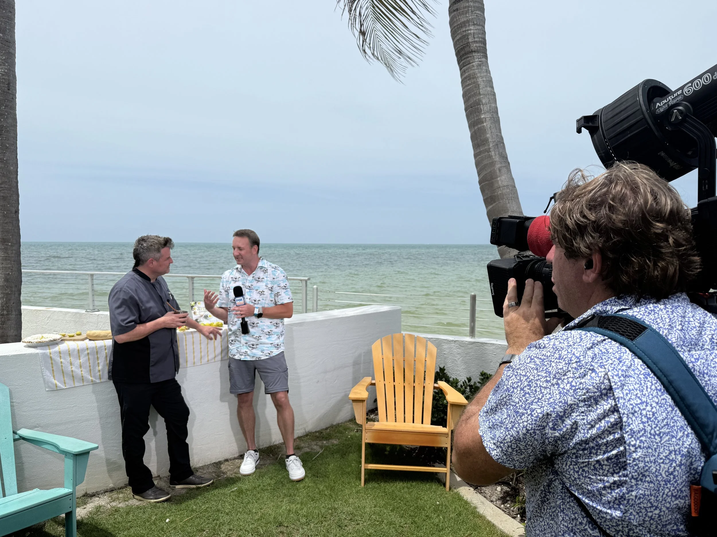  Key Lime Pie expert David Sloan is being interviewed outdoors near the ocean by a camera operator. The interview takes place on a grassy area with palm trees, a white wall, and an Adirondack chair.