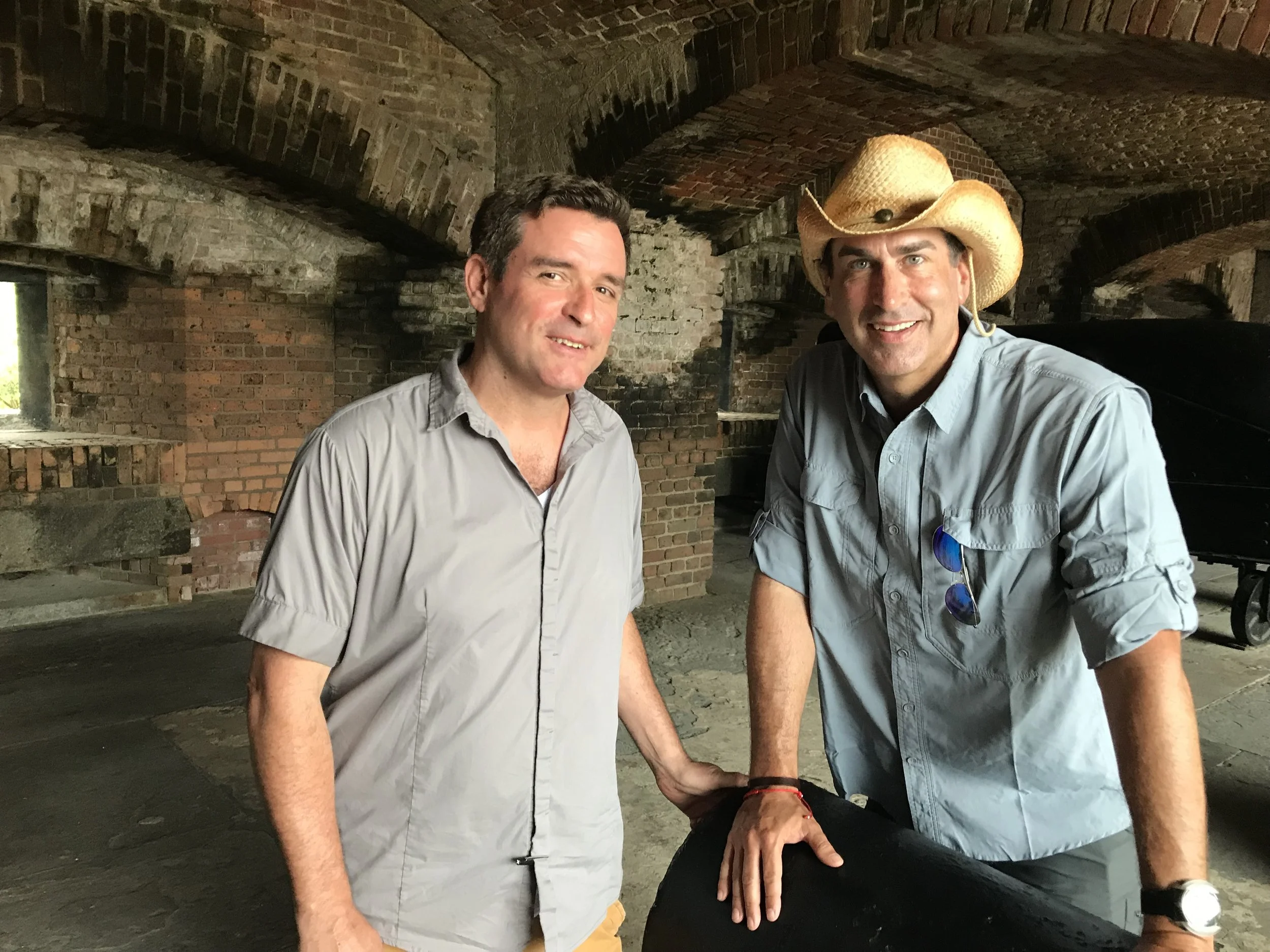 David Sloan and Rob Riggle are smiling inside a brick tunnel at Key West's Fort Zachary Taylor. One wears a cowboy hat, and the other has short dark hair.