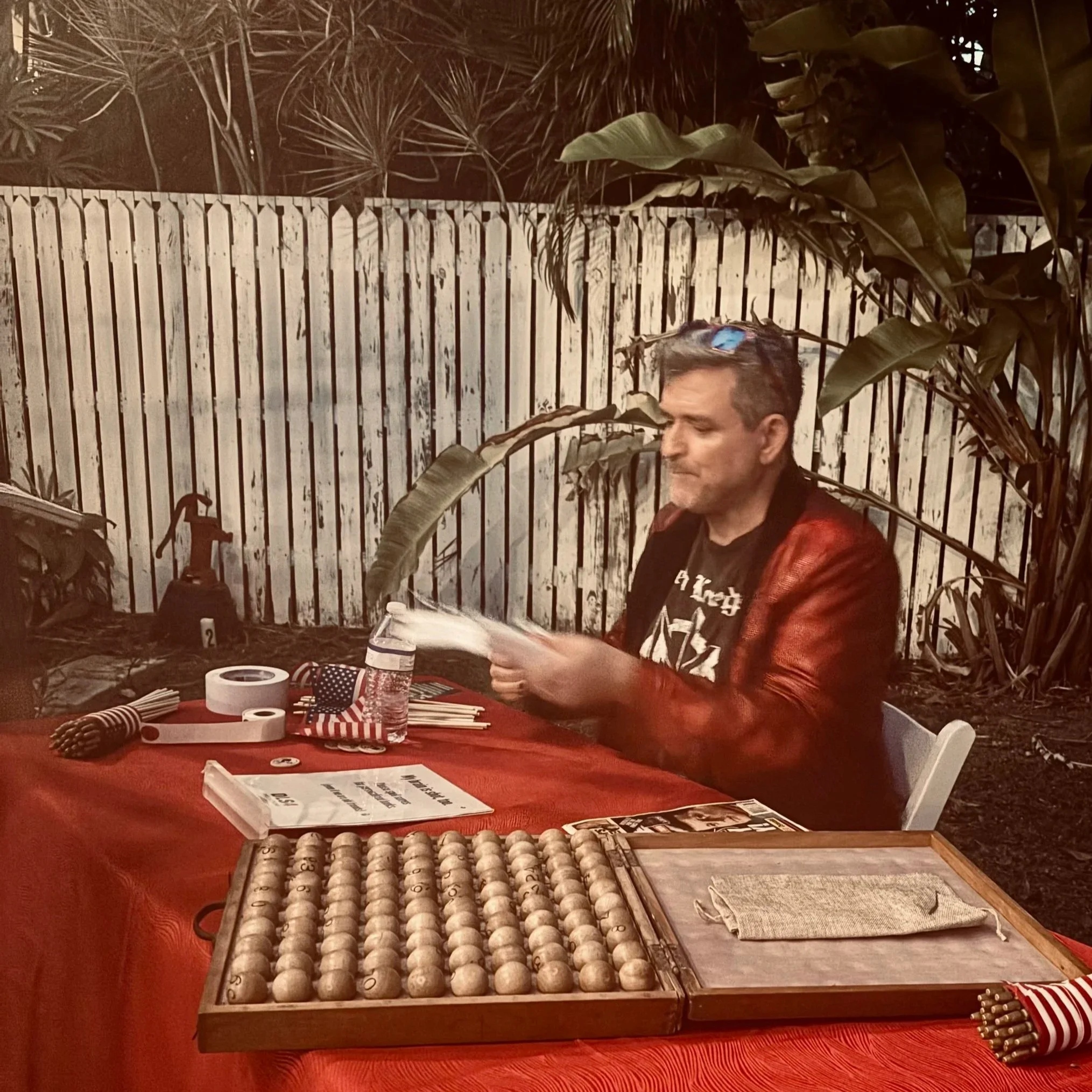 Key West author David L. Sloan is sitting at an outdoor table, signing a book. The table has a red tablecloth, bolita balls, markers, and a bottle of water. In the background is a white wooden fence and some tropical plants.
