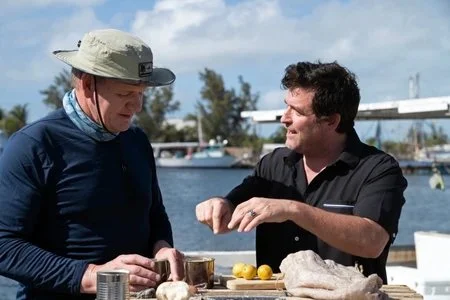 David Sloan and Gordon Ramsay are talking outdoors near water, with boats in the background, preparing Key lime pie on a table with Key limes and utensils.