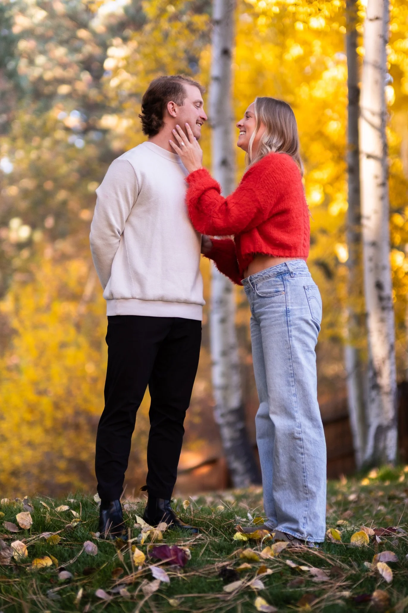 A couple standing outdoors in a forest with autumn leaves, smiling and looking at each other; the woman is touching the man's face gently.
