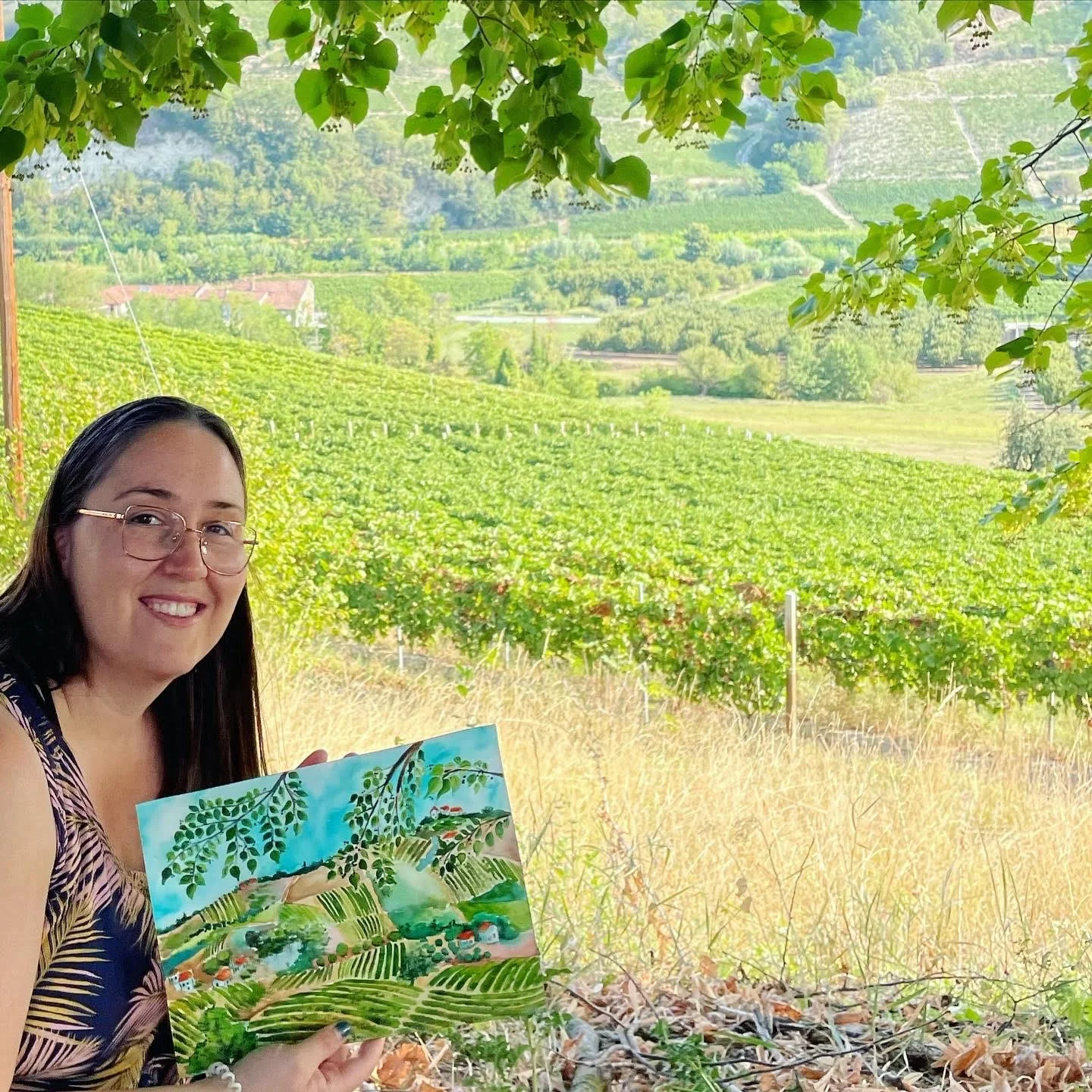 A woman with long dark hair, glasses, and a sleeveless patterned top smiling while holding a colorful painting, outdoors in a vineyard landscape with lush green grapevines, rolling hills, and trees in the background.
