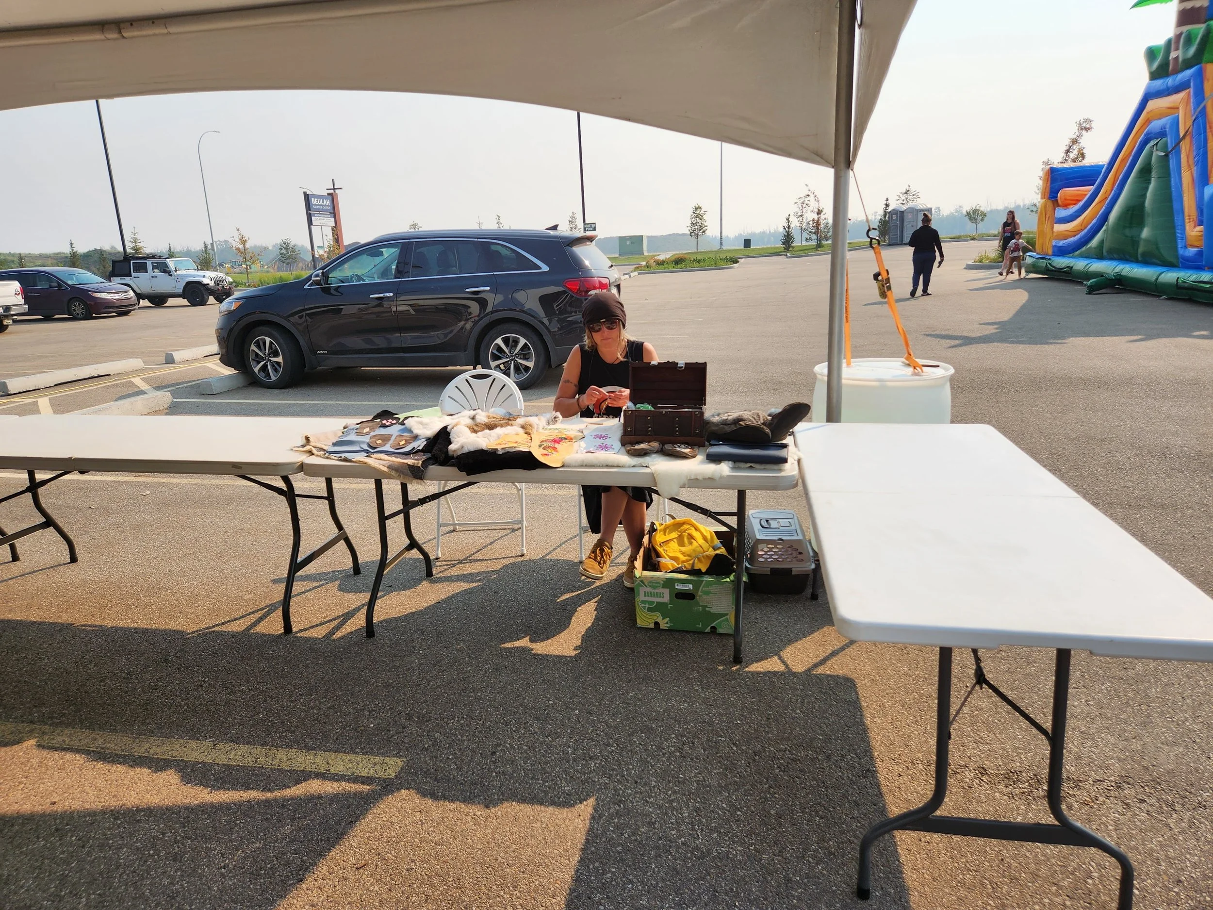 A woman sitting at a table under a canopy at an outdoor market, arranging items for sale, with cars and an inflatable bounce house in the background.