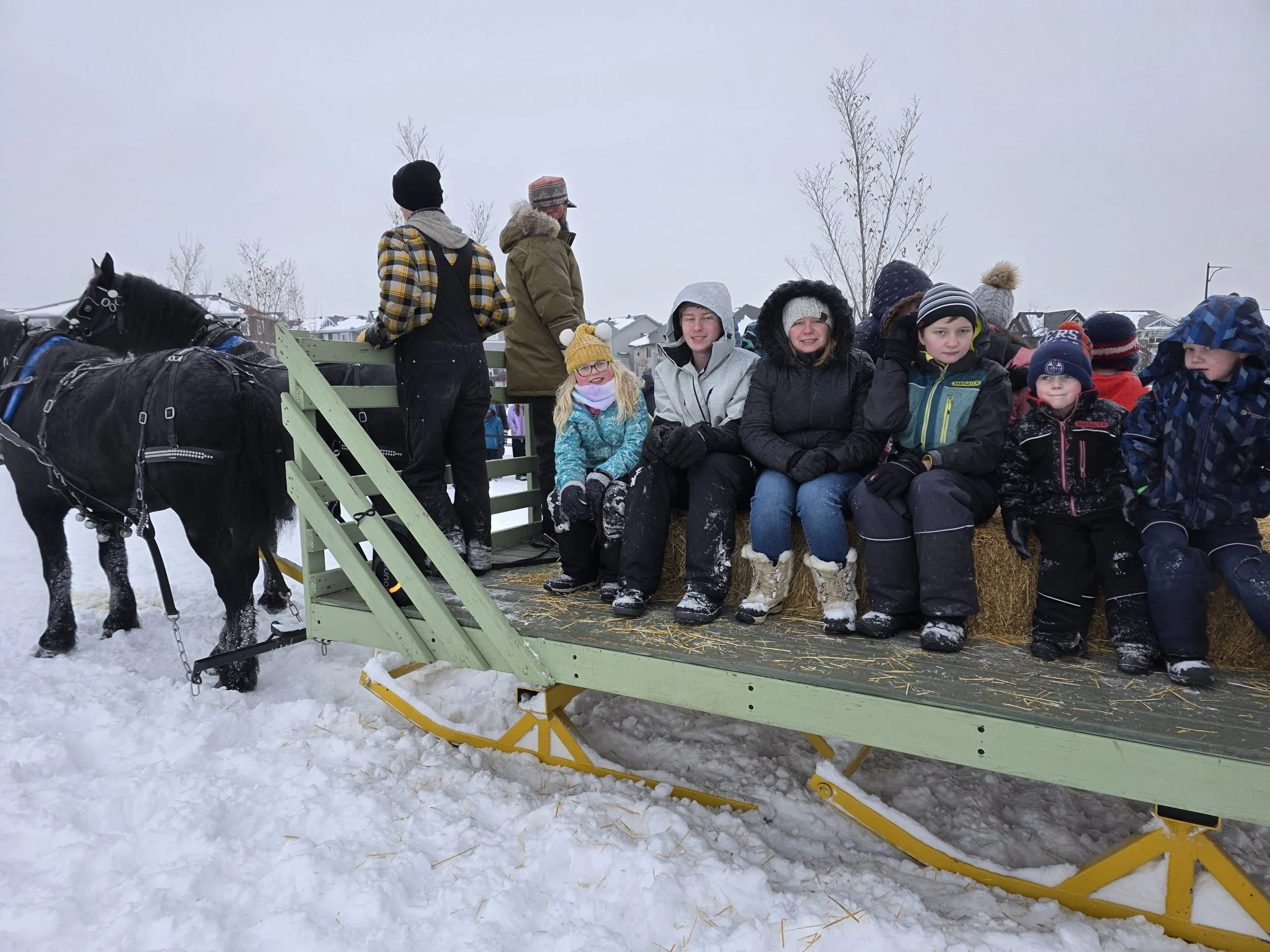 Children and adults sitting on a hayride in snowy weather with a horse-drawn wagon in the background.